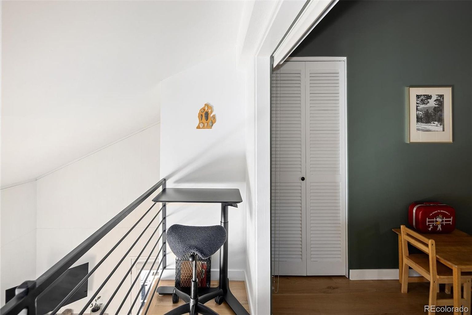 This cozy, multi-functional loft space features a modern standing desk with an ergonomic saddle chair, positioned next to a sleek black metal railing overlooking the lower level. To the right, a dark accent wall provides a sophisticated backdrop for a small wooden desk and a vintage-style red suitcase, while a white louvered closet door adds texture to the room. The combination of light wood flooring and contrasting wall colors creates a bright, contemporary atmosphere perfect for a home workspace.