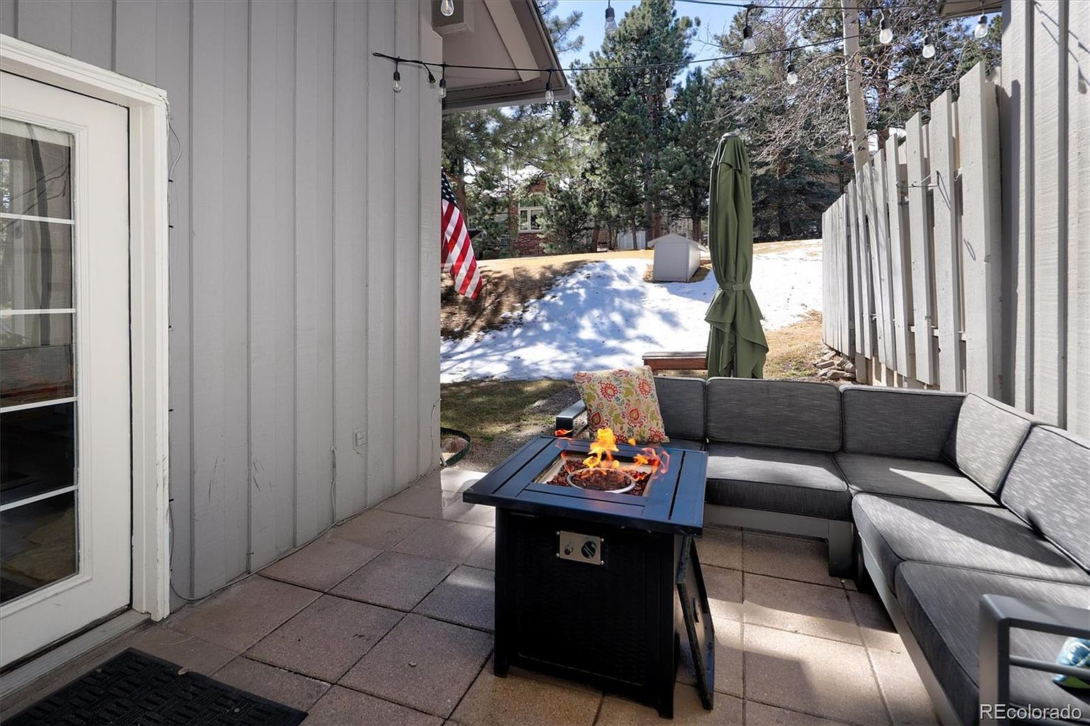 This inviting outdoor patio space features a modern sectional sofa with plush cushions and a central fire pit, creating a cozy atmosphere for relaxation. The area is framed by a light gray exterior wall and a privacy fence, with string lights overhead adding a warm, ambient touch. Beyond the patio, a glimpse of a snowy yard and mature trees provides a serene, seasonal backdrop.