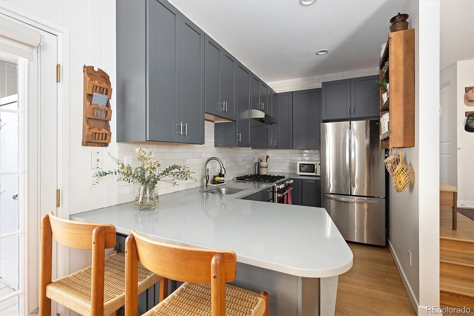 This modern kitchen features sleek, dark grey cabinetry paired with a light-colored quartz countertop and a white subway tile backsplash. The space includes stainless steel appliances, a peninsula with two wooden bar stools, and warm wood-tone flooring, creating a cozy yet contemporary atmosphere. The perspective is from the dining area looking into the kitchen, highlighting the efficient layout and functional design.