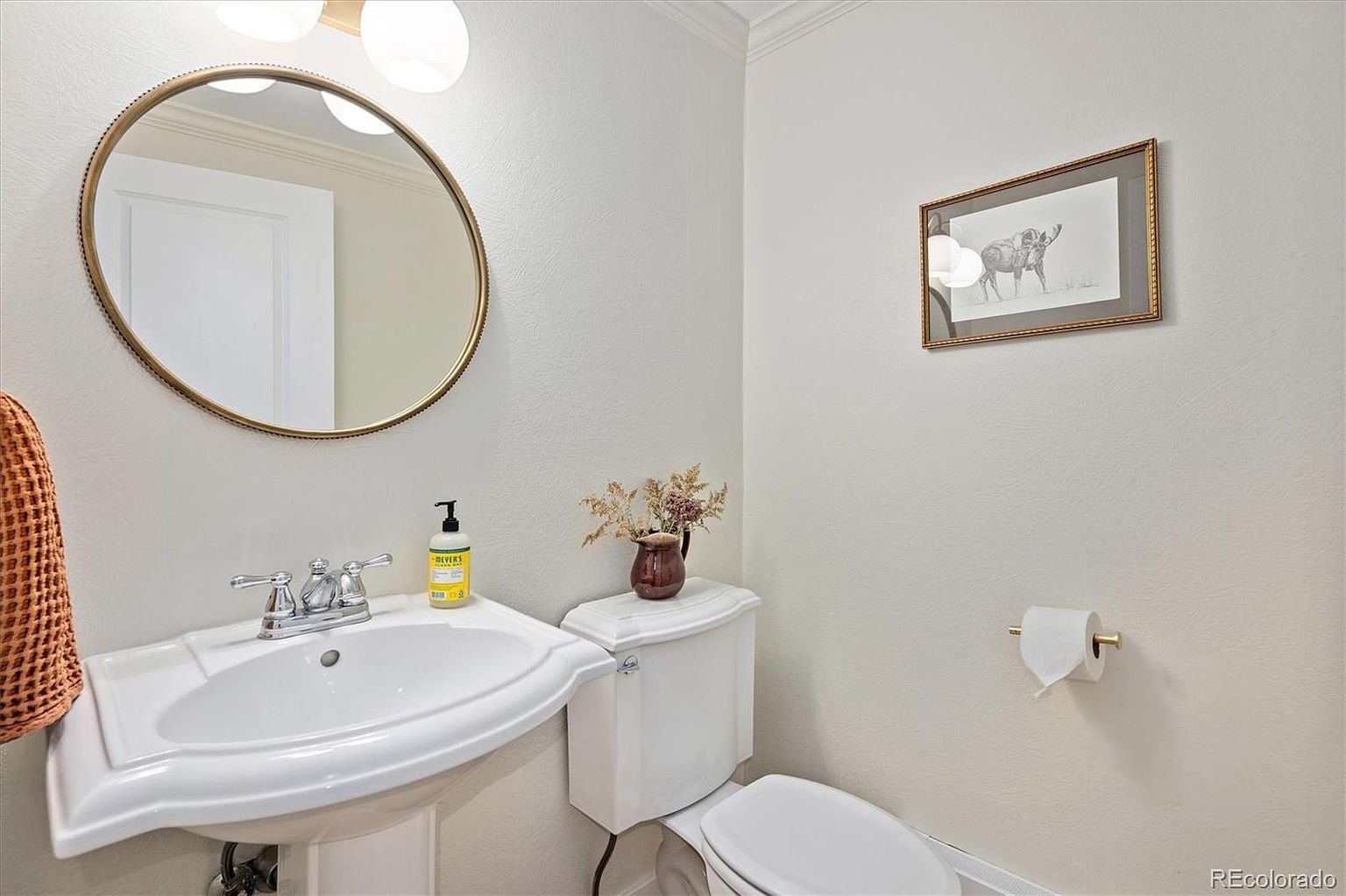 This compact powder room features a classic pedestal sink with a polished chrome faucet and a round, gold-rimmed mirror. The space is decorated with a framed moose print on the wall and a small vase of dried flowers atop the toilet tank, creating a cozy and rustic aesthetic. The neutral wall color and simple fixtures provide a clean, inviting atmosphere for guests.