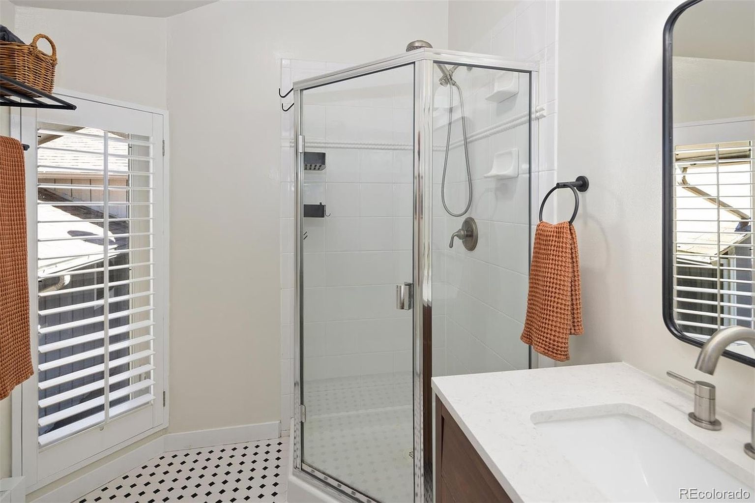 This bright and clean bathroom features a corner glass-enclosed shower with white subway tile and a modern vanity with a white stone countertop. A black-framed mirror hangs above the sink, complementing the black hardware and towel ring, while the floor is finished with a classic black-and-white patterned tile. The space is well-lit by natural light streaming through plantation shutters, creating a crisp and inviting atmosphere.