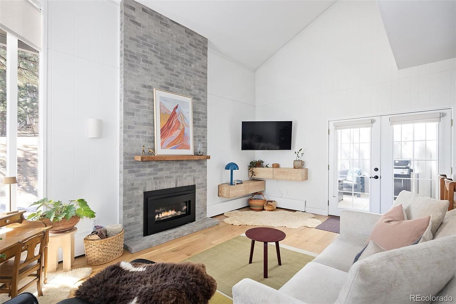 This bright and airy living room features a striking floor-to-ceiling gray brick fireplace as its focal point, complemented by a natural wood mantel and a modern gas insert. The space is anchored by light hardwood floors, a neutral sofa, and floating wood shelving, creating a warm, contemporary aesthetic. Large windows and glass doors flood the room with natural light, offering a seamless connection to the outdoors.