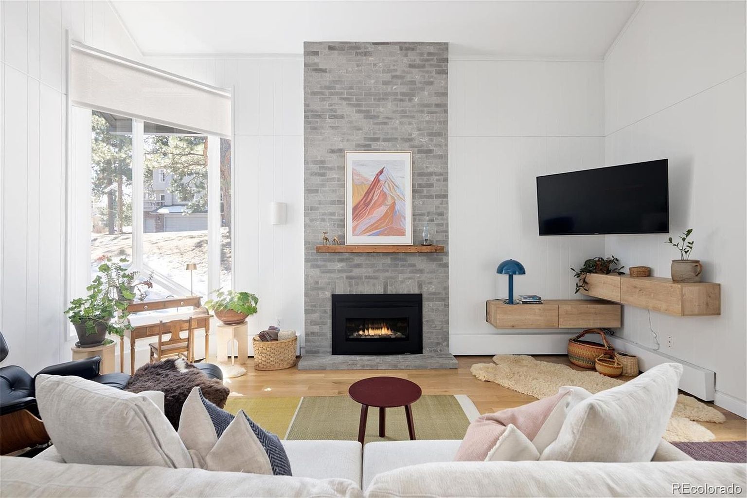 This bright and airy living room features a striking grey brick fireplace as its central focal point, complemented by a warm wood mantel and a large abstract mountain print. The space is furnished with a comfortable white sofa, a modern circular coffee table, and floating light-wood media consoles, all set against clean white walls and light hardwood floors. Large windows on the left invite natural light, creating a cozy, contemporary atmosphere perfect for relaxation.