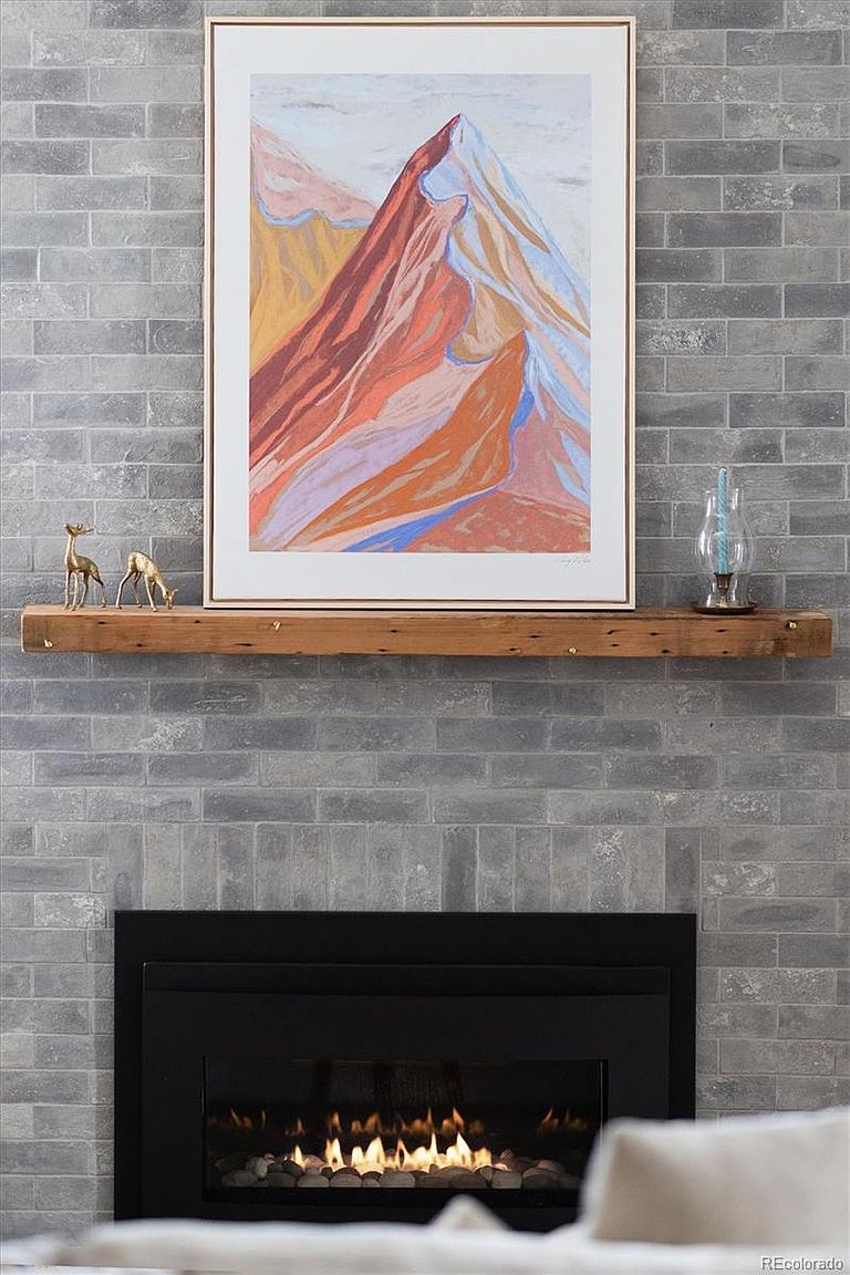 This cozy living room focal point features a modern fireplace with a sleek black insert set against a textured gray brick wall. A rustic, reclaimed wood mantelpiece holds a framed abstract mountain landscape painting, flanked by two small brass deer figurines and a glass candle holder. The composition is centered and symmetrical, creating a warm, inviting atmosphere perfect for a home listing.
