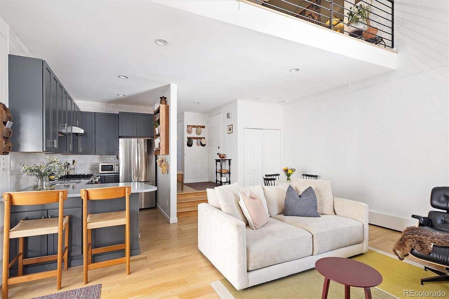 This open-concept living space features a bright, modern aesthetic with light hardwood floors and a neutral color palette. The area seamlessly connects a cozy living room, furnished with a plush beige sofa and a classic lounge chair, to a sleek kitchen with dark grey cabinetry and a breakfast bar. The high ceilings and lofted overlook create an airy, spacious feel, making it an inviting and functional home layout.