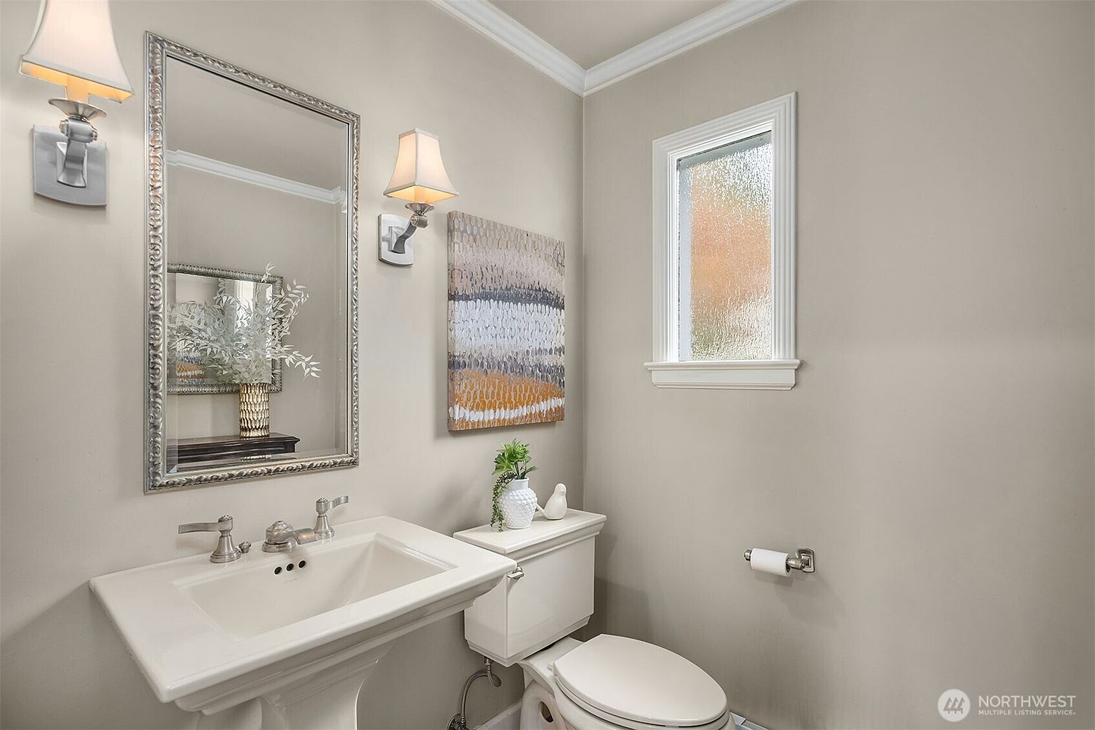 This is a well-lit guest bathroom featuring a pedestal sink with a decorative mirror, a toilet with a white ceramic bird figurine on top, and a small window providing natural light. The walls are painted in a neutral tone, complemented by white trim and fixtures, creating a clean and inviting space. An abstract painting hangs on the wall between the mirror and the window.