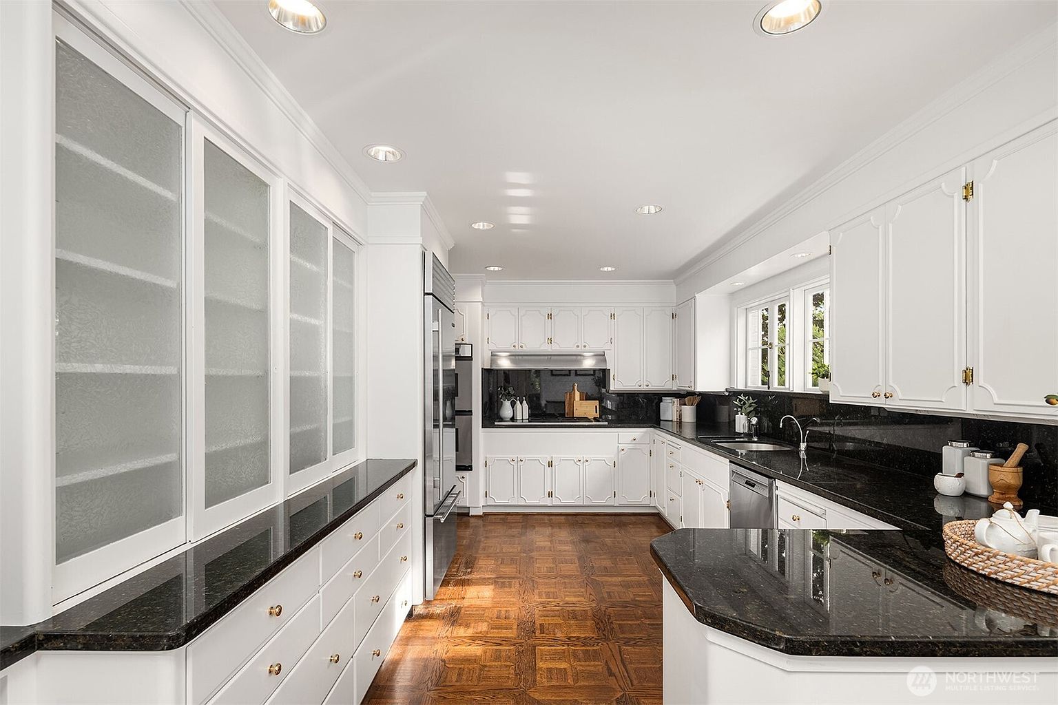 This is a well-lit kitchen featuring white cabinetry, black granite countertops, and hardwood flooring. The kitchen includes a large pantry with frosted glass doors, stainless steel appliances, and a window providing natural light. The overall impression is a clean and classic design.