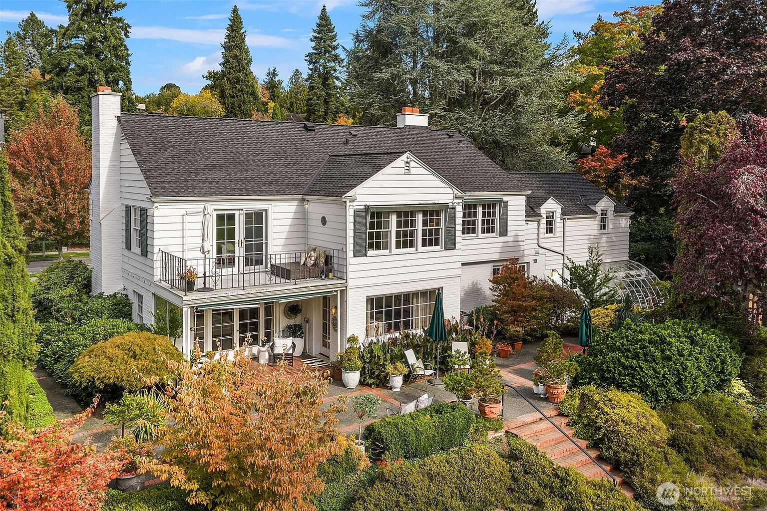 This aerial view showcases a grand white house with a dark gray roof, surrounded by lush greenery and mature trees. The property features a well-manicured garden with brick pathways and multiple outdoor seating areas, including a balcony and a patio. The overall impression is one of elegance and tranquility, highlighting the home's curb appeal and outdoor living spaces.