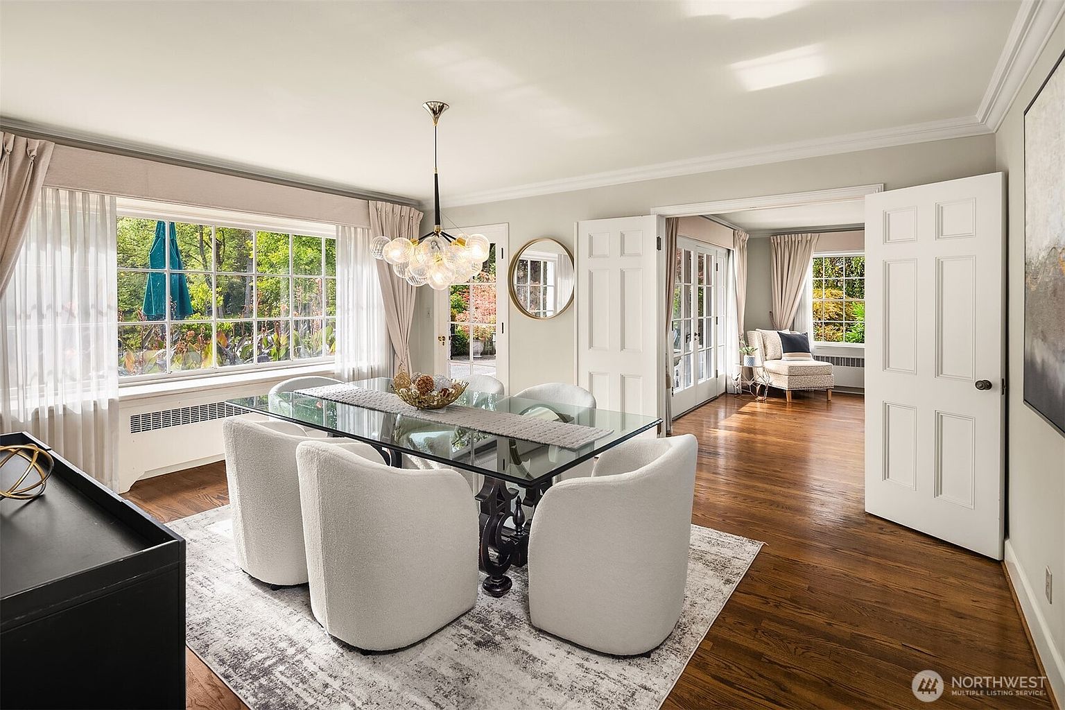 This is an interior shot of a dining room featuring a glass-top table with a dark base, surrounded by four white upholstered chairs. A modern chandelier hangs above the table, and a large window provides natural light. The room is decorated in neutral tones with hardwood floors and a patterned rug, creating an elegant and inviting atmosphere.
