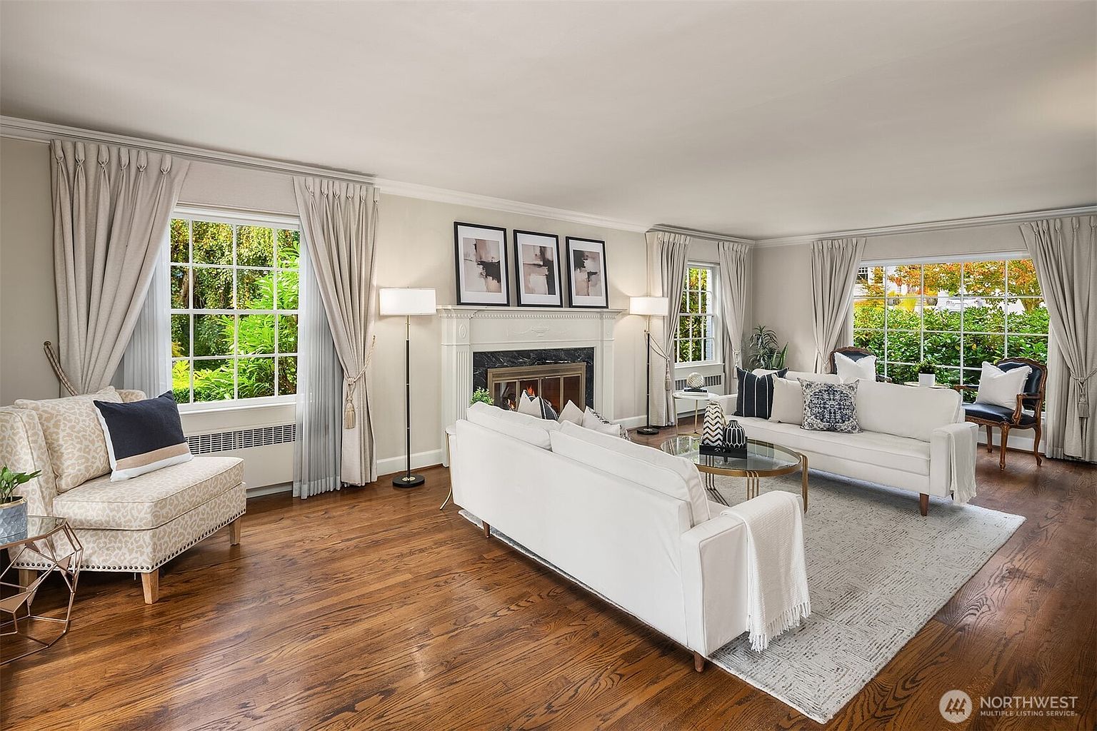 This is a bright and inviting living room featuring hardwood floors, neutral-toned walls, and large windows that provide ample natural light. The room is furnished with two white sofas, a patterned armchair, and a fireplace with decorative artwork above. The overall impression is one of comfort and elegance, perfect for relaxing or entertaining.