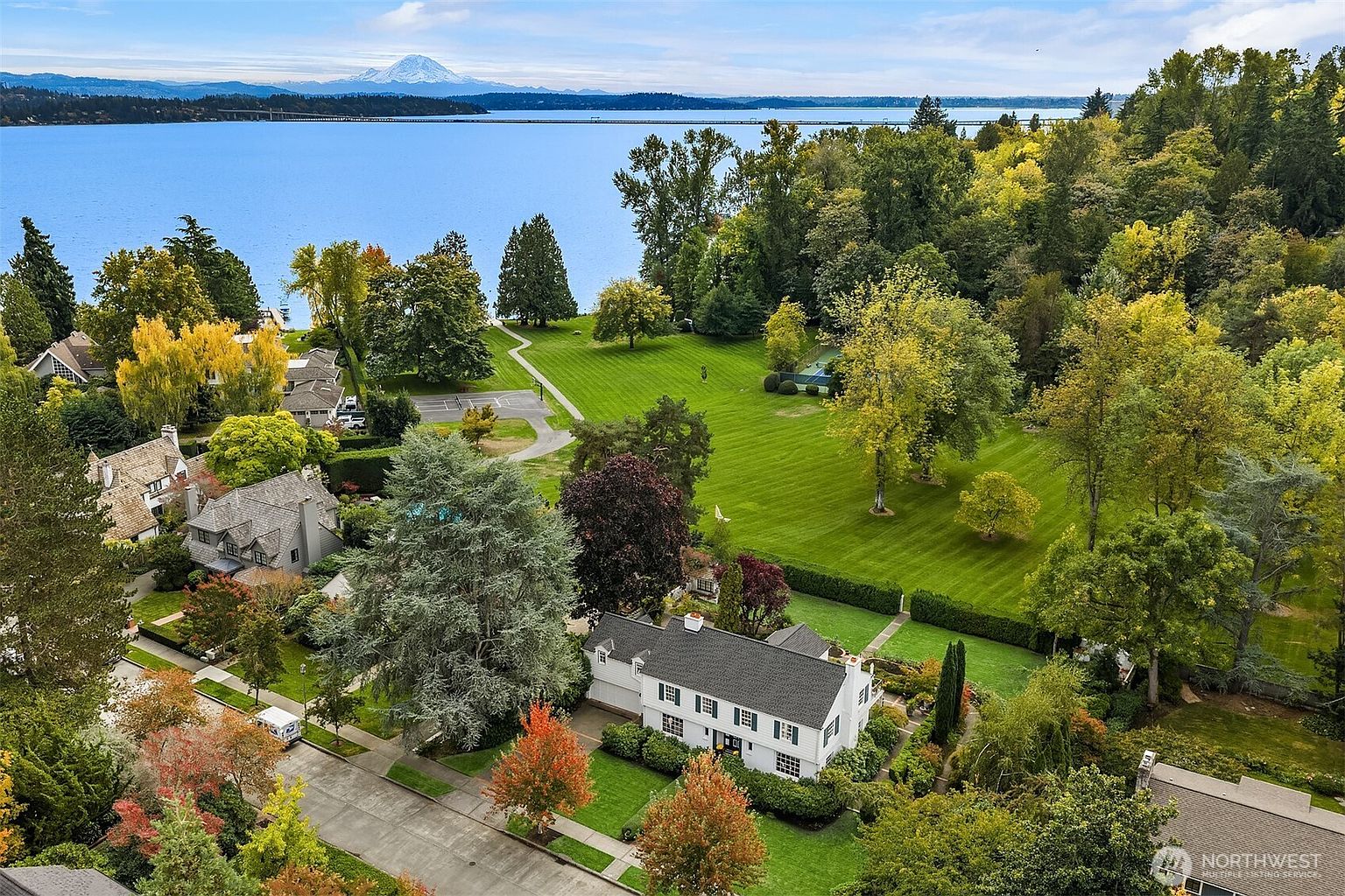 This aerial shot showcases a beautiful property with a well-maintained white house featuring a dark roof and green shutters, surrounded by lush green lawns and mature trees displaying autumn colors. The property extends to a large grassy area bordering a lake, with a mountain visible in the distance, creating a serene and picturesque setting. The view emphasizes the property's size, landscaping, and desirable location.