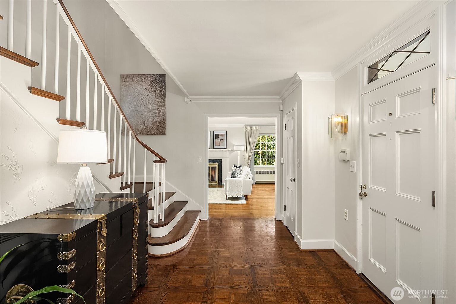 This is an interior shot of a home's entryway and staircase. The foyer features dark wood parquet flooring, a white front door with decorative glass, and a staircase with white railings and wood treads. A glimpse into the living room reveals a fireplace and a bright, inviting space, enhancing the home's appeal.