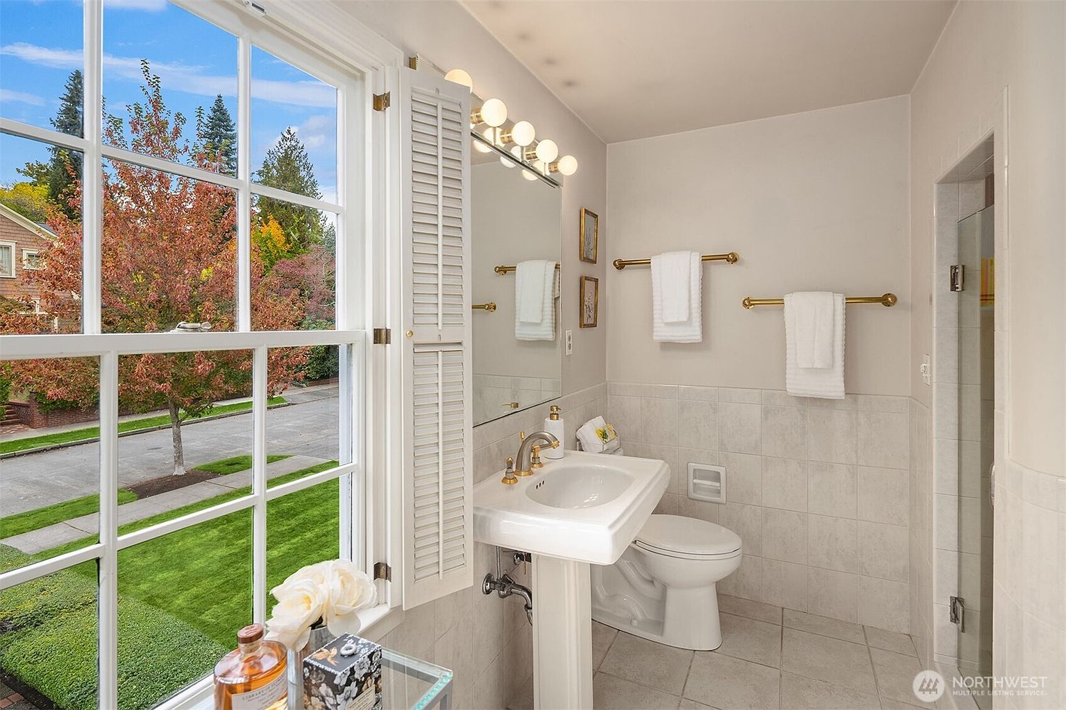 This is a well-lit bathroom featuring a pedestal sink with gold fixtures, a toilet, and a shower with glass doors. The walls are tiled, and there are gold towel racks with white towels. A window with white shutters offers a view of a tree with autumn foliage, adding natural light and a touch of nature to the space.