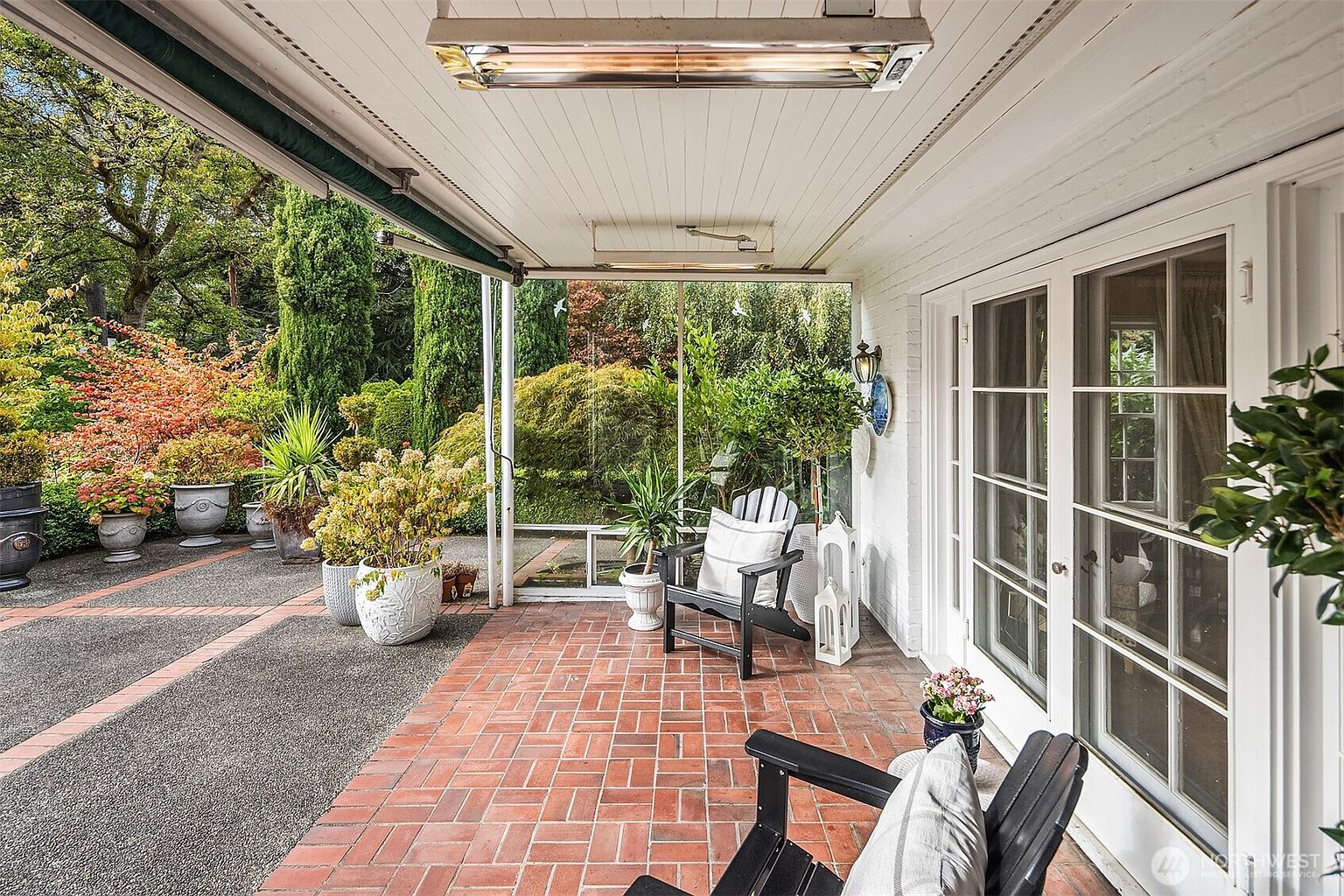 This image showcases a charming outdoor patio area, featuring a brick floor and a covered ceiling with a heater. Comfortable black Adirondack chairs with white cushions invite relaxation, while potted plants add a touch of greenery. The patio offers a view of a well-maintained garden, creating a serene and inviting atmosphere.