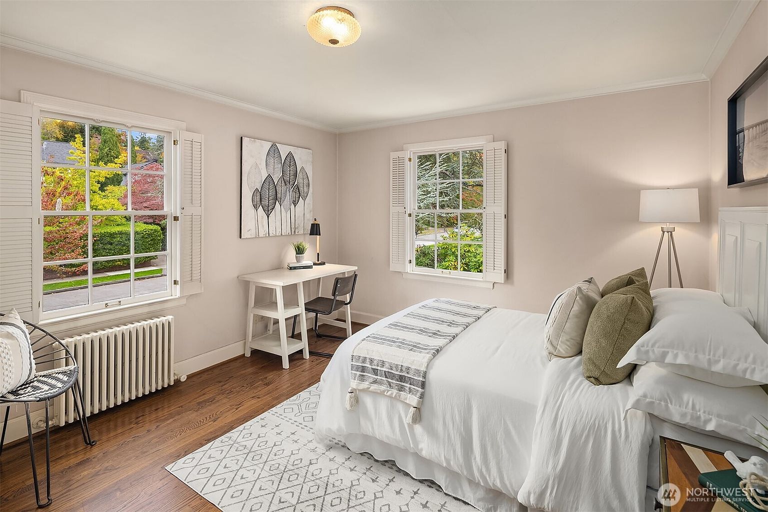 This is a cozy bedroom featuring hardwood floors, two windows with white shutters, and a white bed with neutral-toned pillows and a throw blanket. A white desk with a black chair sits near one window, and a radiator is positioned under the other. The room has a warm and inviting atmosphere, perfect for a guest room.