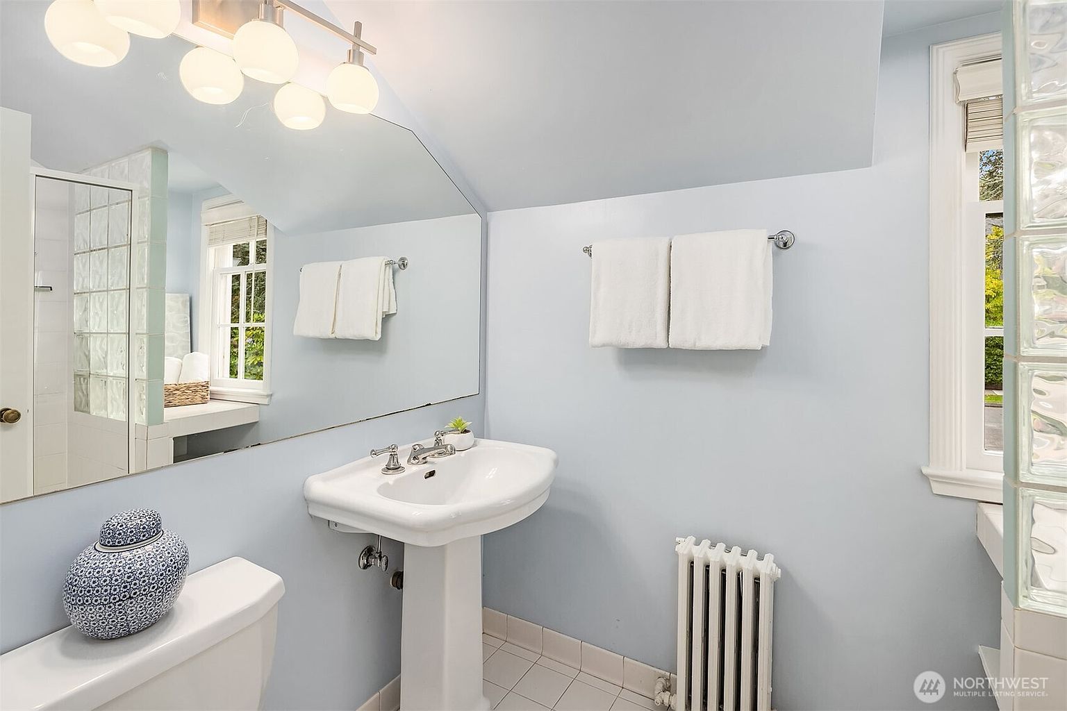 This is a bright and clean bathroom featuring a pedestal sink, a large mirror, and a classic radiator. The walls are painted a soft blue, and the room is well-lit by both overhead lighting and natural light from a window with glass blocks. A decorative blue and white jar sits on the toilet tank, adding a touch of elegance.