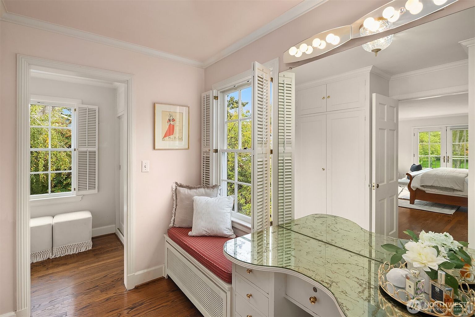 This is a charming primary bathroom featuring a built-in window seat with red cushion and decorative pillows, a vanity with a mirrored top, and white cabinetry. The room is painted in a soft pink hue, complemented by white trim and shutters. Natural light streams through the windows, creating a bright and inviting space.