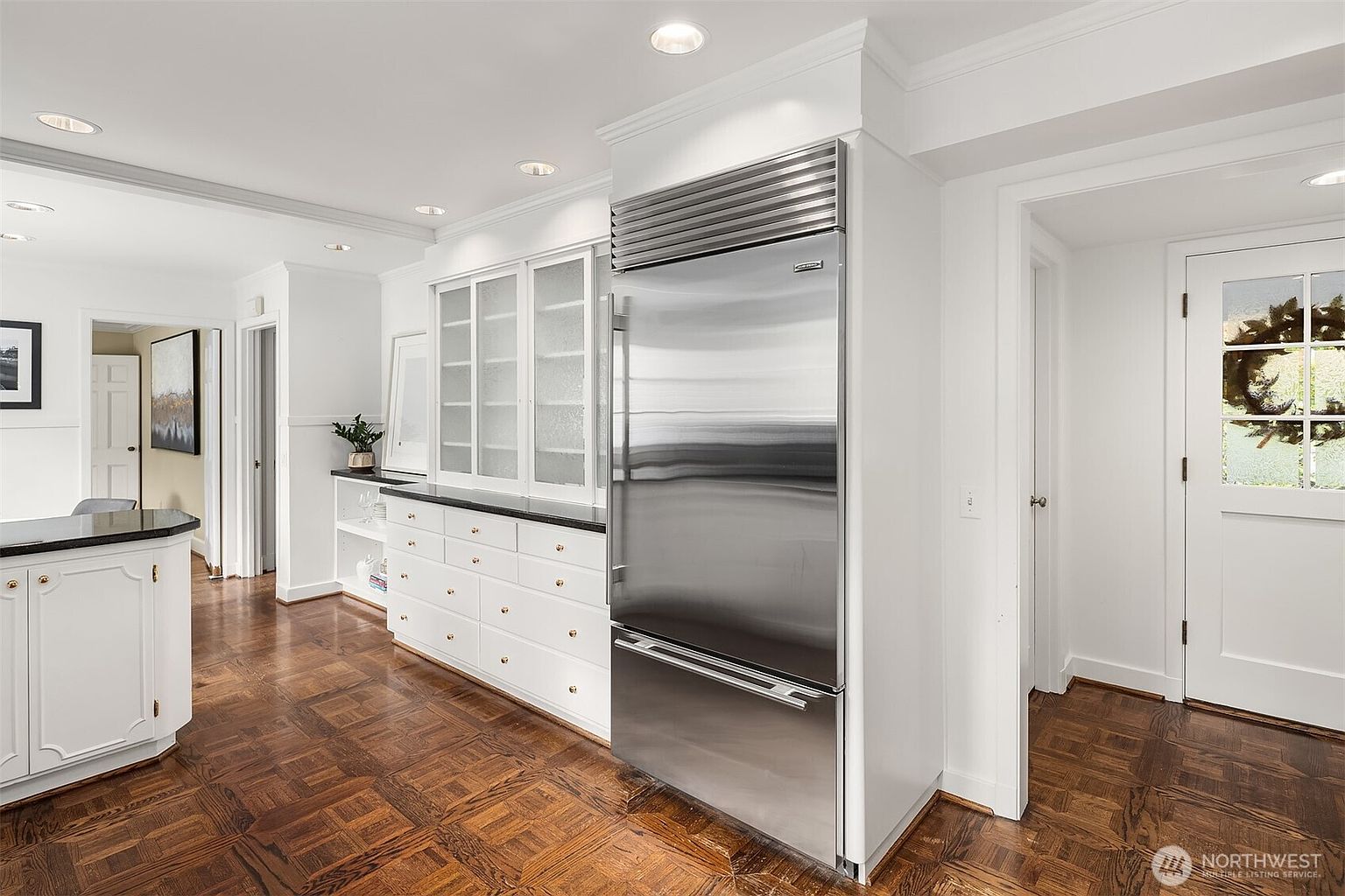 This interior shot showcases a well-lit kitchen area with a classic design. The kitchen features white cabinetry with gold hardware, a black countertop, and a stainless steel refrigerator. The parquet wood flooring adds warmth and character to the space, while the white walls and trim create a bright and airy atmosphere.