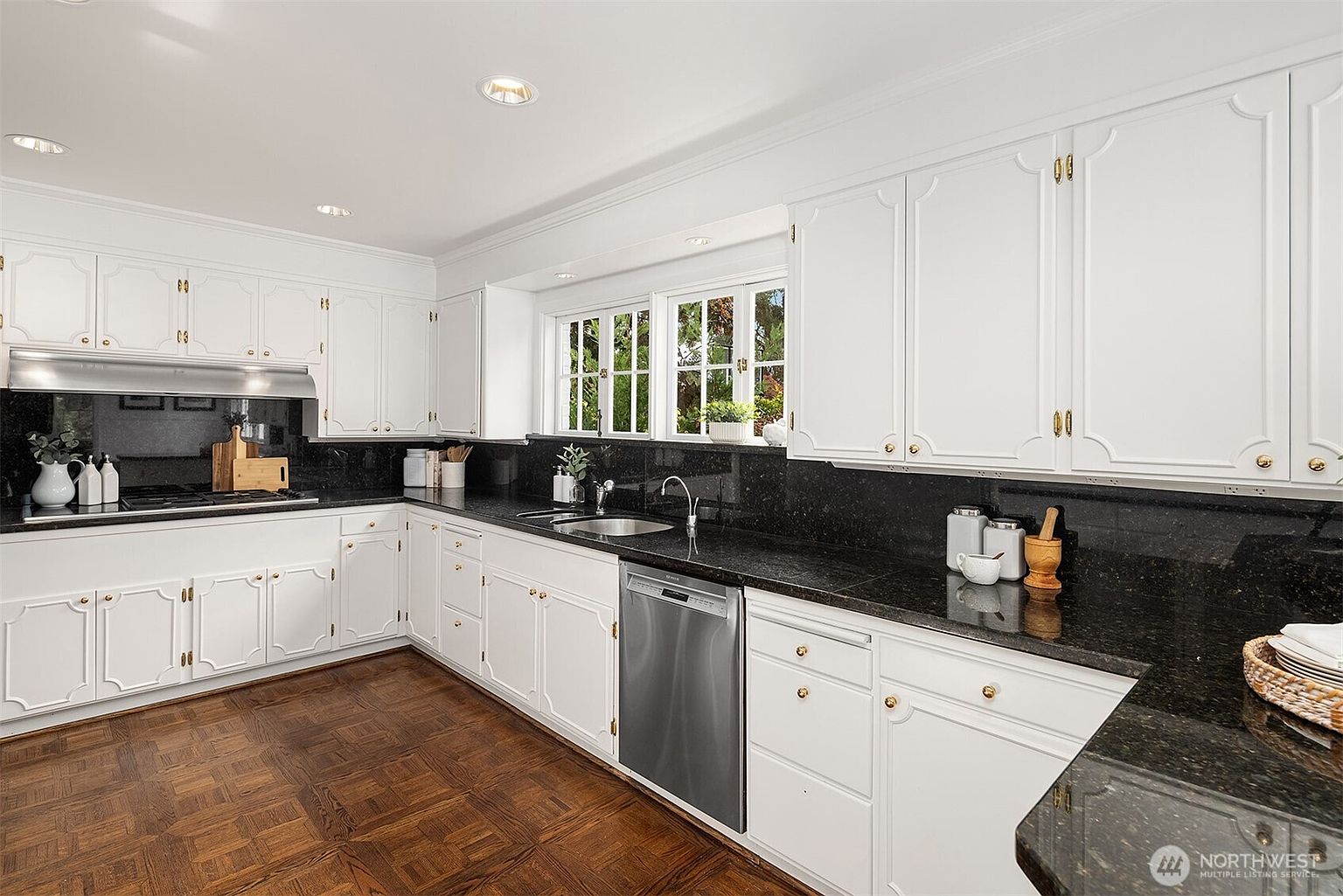This is a well-lit kitchen featuring white cabinetry with gold hardware and dark granite countertops. The kitchen includes stainless steel appliances and a window above the sink that provides natural light. The flooring is a patterned wood, adding warmth to the space.