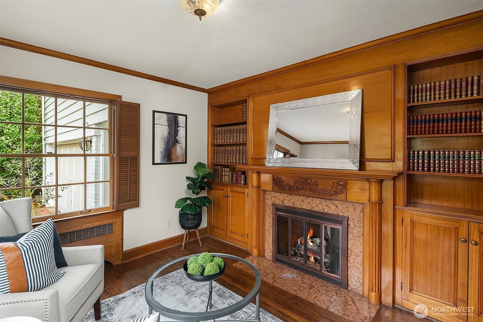This is an interior shot of a living room featuring a traditional style with wood paneling and built-in bookshelves flanking a fireplace. The fireplace has a decorative mantel and a mirror above it. A window with shutters provides natural light, and a round coffee table sits in front of a chair, adding to the cozy and inviting atmosphere of the room.