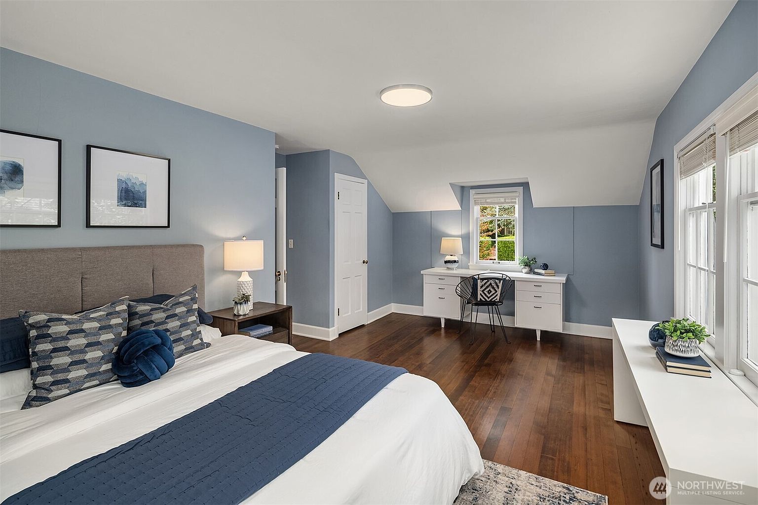 This is a well-lit bedroom featuring a bed with blue and white bedding, a gray upholstered headboard, and two framed pictures on the wall. The room has a desk area with a chair and a window, and the walls are painted in a calming blue tone with white trim near the ceiling. The hardwood floors add warmth to the space.