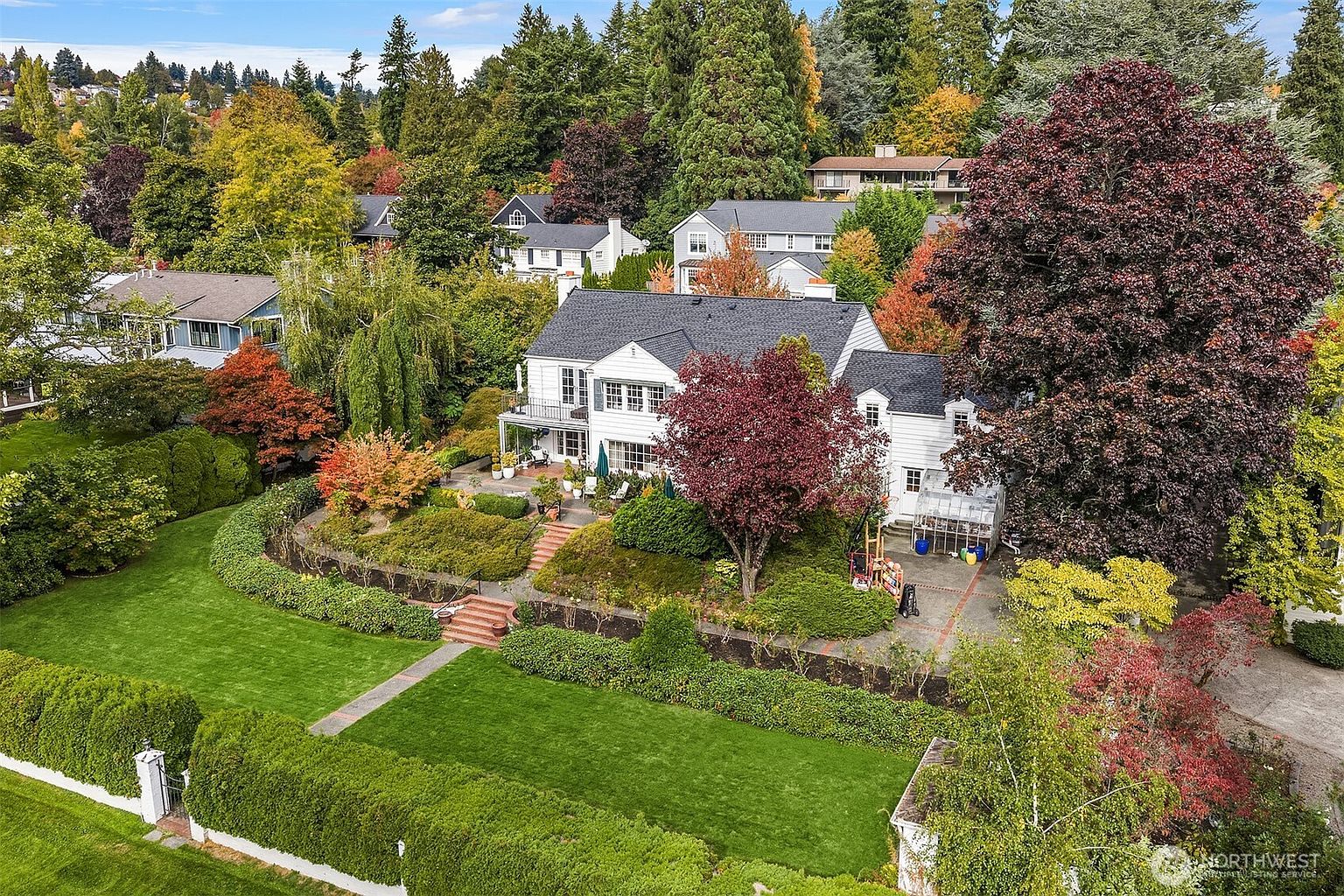 This aerial view showcases a beautifully landscaped property with a large white house featuring a dark roof. The yard boasts lush green lawns, manicured hedges, and mature trees displaying vibrant fall colors. A stone pathway leads to the house, enhancing the property's curb appeal and inviting atmosphere.
