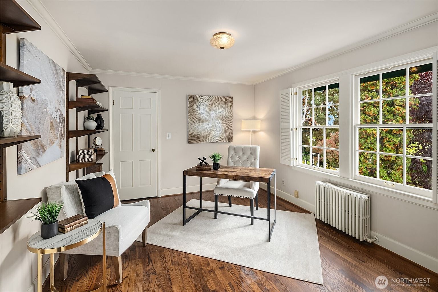 This is a well-lit home office featuring hardwood floors, a neutral color palette, and natural light from two large windows. The room is furnished with a wooden desk, a tufted chair, a small seating area with a side table, and built-in shelving. The overall impression is a comfortable and functional workspace.