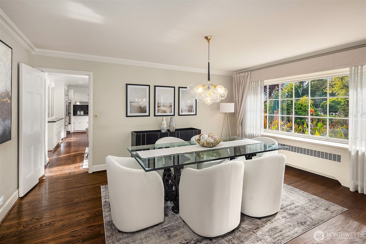 This is an interior shot of a dining room featuring a glass-top table with a dark, ornate base, surrounded by four white upholstered chairs. A modern chandelier hangs above the table, and a black sideboard with framed artwork sits against the wall. The room is well-lit with natural light from a large window, creating a bright and inviting atmosphere.