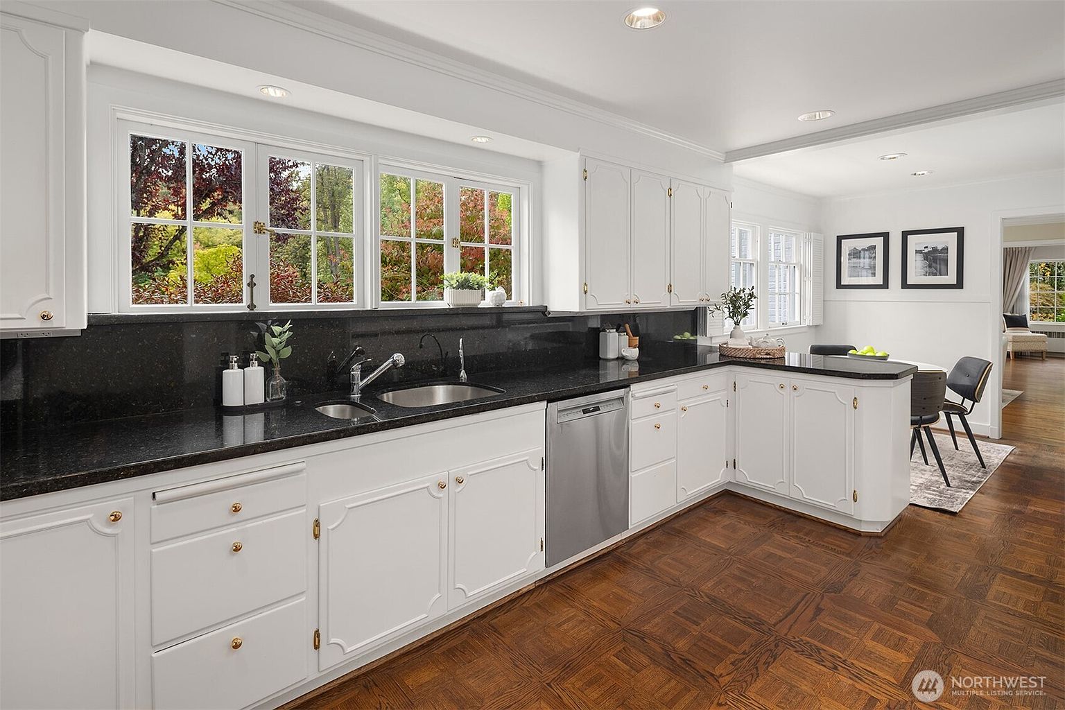 This is a well-lit kitchen featuring white cabinetry with gold hardware and black granite countertops. A stainless steel dishwasher is visible, and a large window provides natural light and a view of the outdoors. The flooring is a classic parquet pattern, adding warmth to the space.