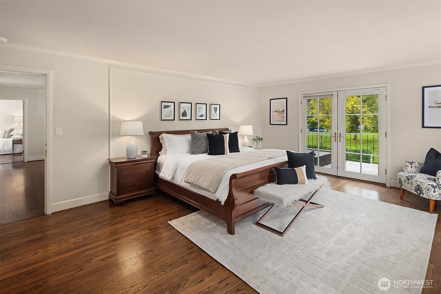 This is a well-lit primary bedroom featuring a large wooden sleigh bed with neutral-toned bedding and decorative pillows. The room has hardwood floors, white walls with molding, and French doors leading to the outside. A nightstand with a lamp sits beside the bed, and artwork adorns the wall above the headboard, creating a serene and inviting atmosphere.