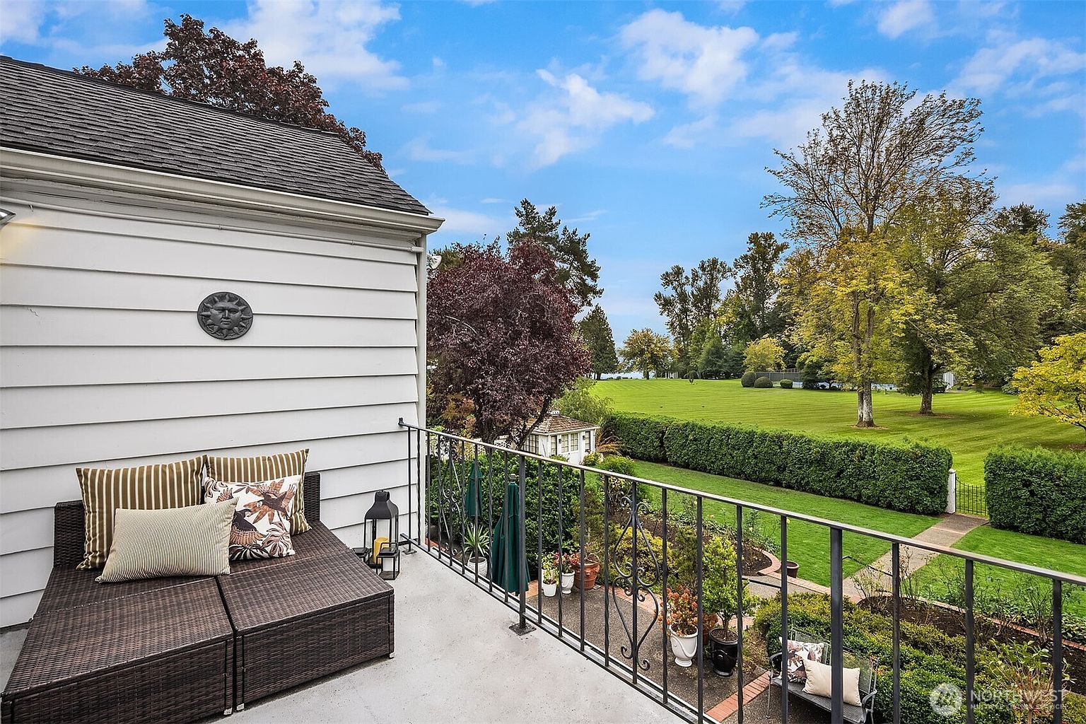 This image showcases a charming balcony with a cozy outdoor seating area, featuring a wicker-style daybed adorned with decorative pillows. The balcony overlooks a meticulously maintained garden and expansive lawn, offering a serene view of the surrounding landscape. The scene evokes a sense of relaxation and outdoor living.