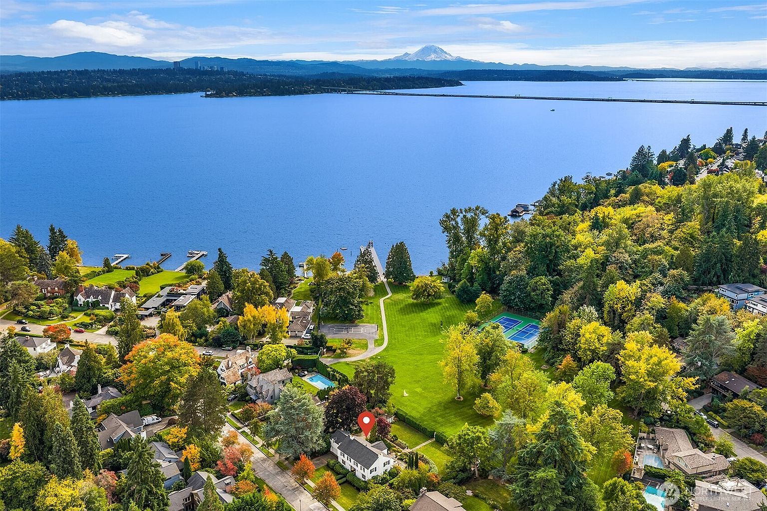 This aerial shot showcases a beautiful property with a well-manicured lawn, mature trees, and a charming house with a dark roof. A tennis court and swimming pool add to the appeal, while the backdrop of a large lake and distant mountains creates a stunning vista. The overall impression is one of luxury and tranquility.