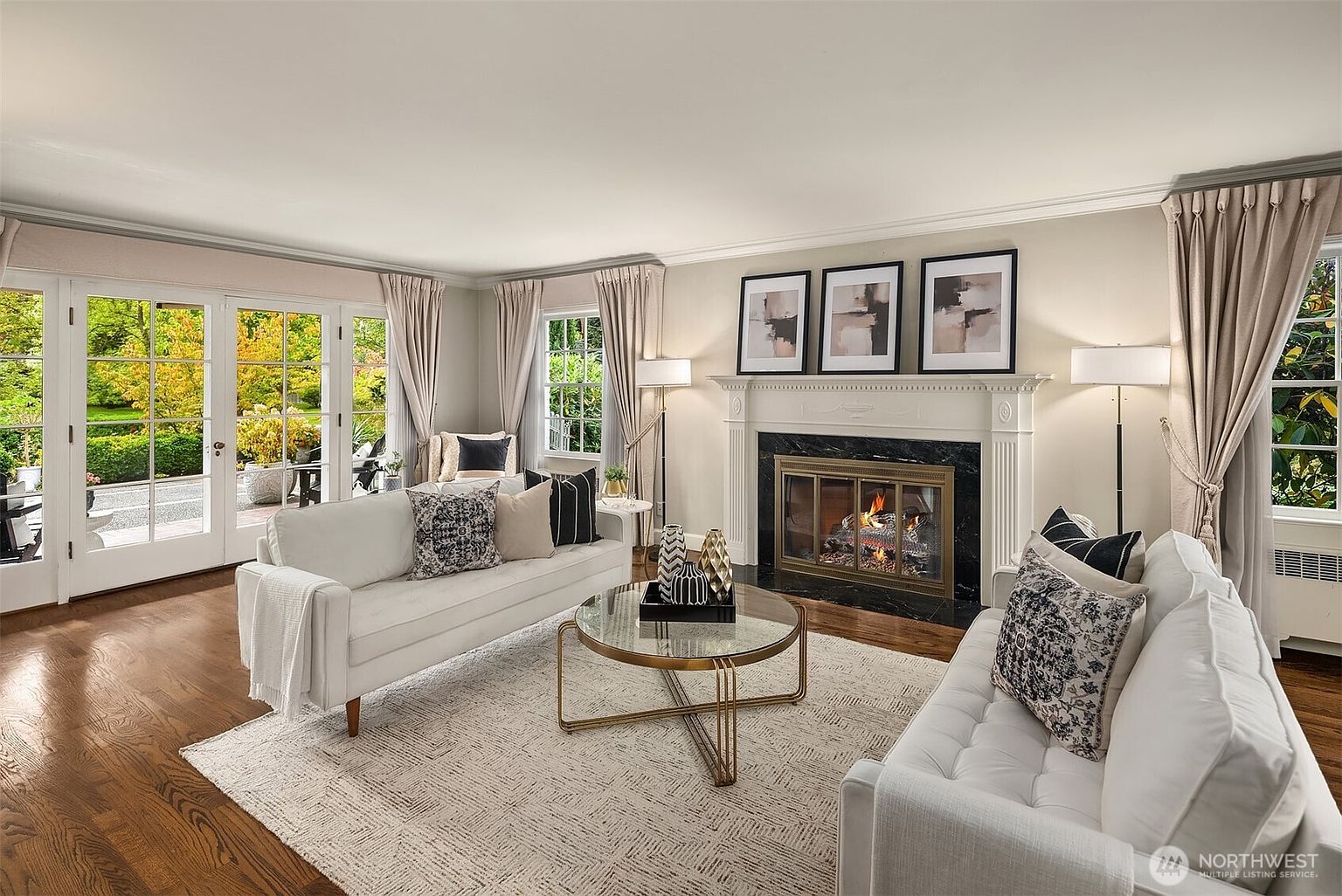 This is a well-lit living room featuring hardwood floors, a fireplace with a black marble surround, and neutral-toned walls. The room is furnished with two white sofas, decorative pillows, and a round glass coffee table. Large windows and French doors provide ample natural light and views of the exterior landscape, enhancing the room's inviting atmosphere.