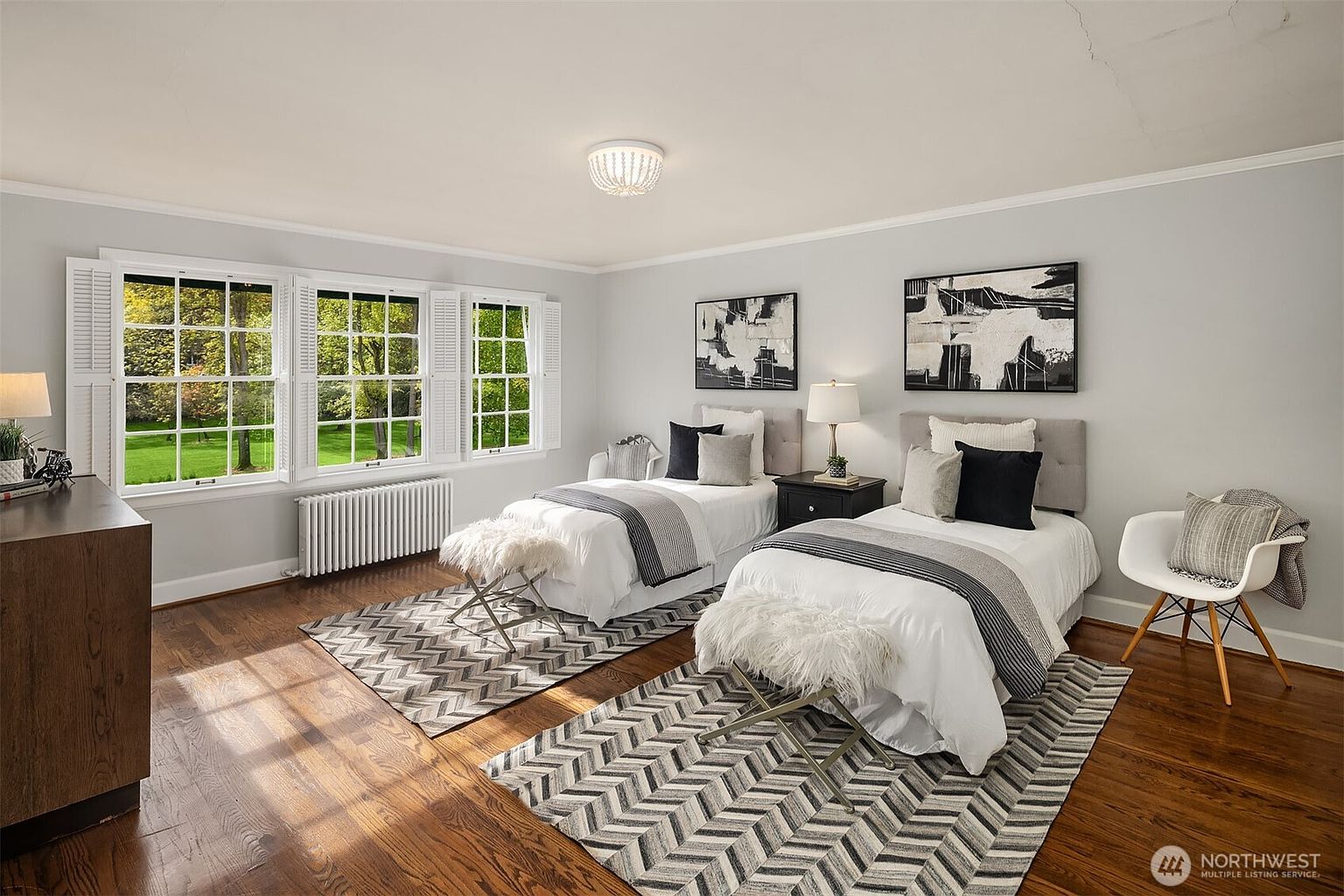 This is a bright and inviting guest bedroom featuring two twin beds, each adorned with neutral-toned bedding and decorative pillows. The room is well-lit by natural light streaming through the windows, complemented by stylish artwork and a modern chandelier. The hardwood floors and patterned rugs add warmth and texture to the space, creating a comfortable and aesthetically pleasing environment.