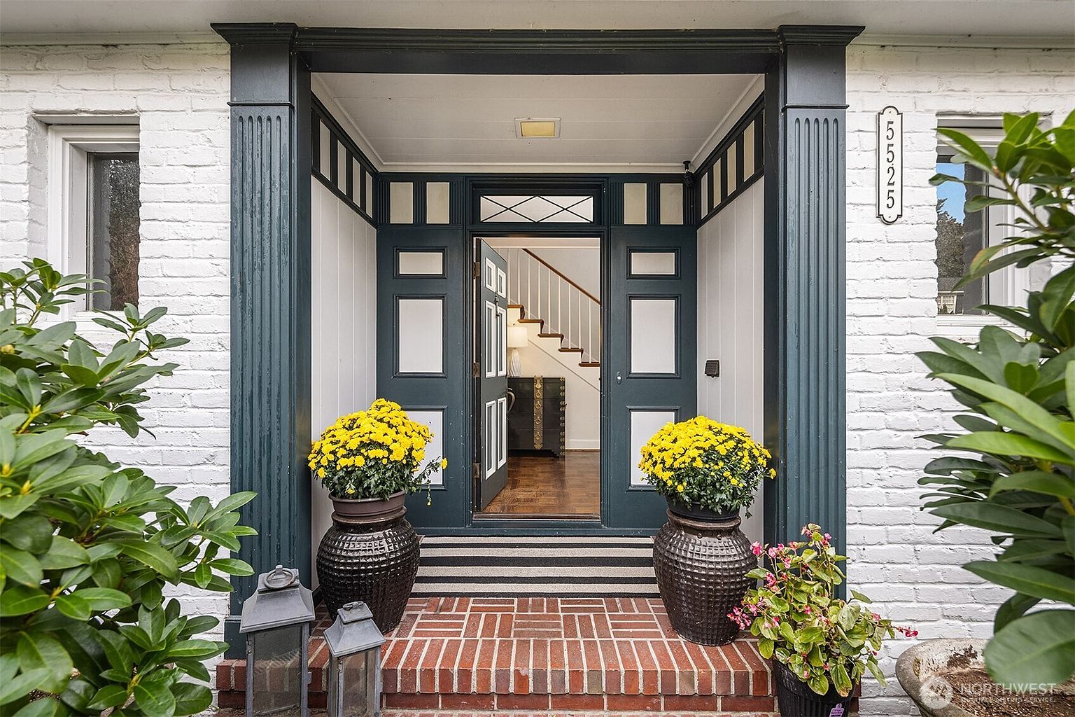 This is a welcoming entryway to a home, featuring a brick stoop with a striped rug leading to a dark blue front door that is slightly ajar. Large potted yellow flowers flank the entrance, adding a pop of color. The exterior is painted white brick, and the house number is visible on the right side of the door frame.