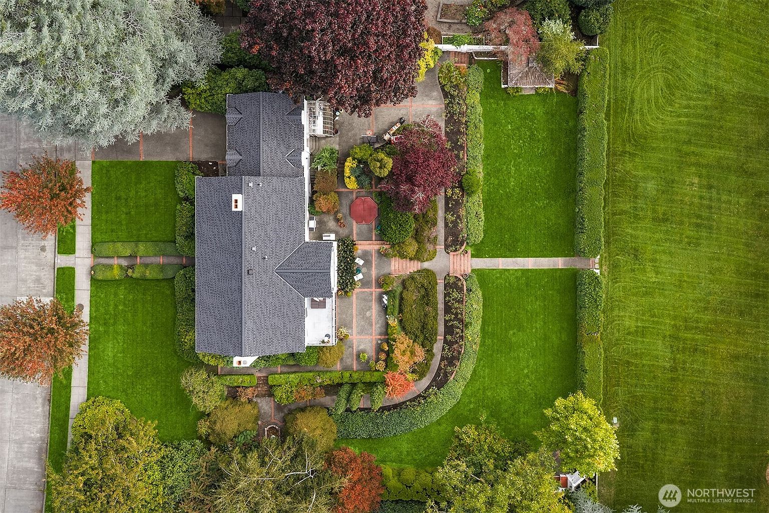 This aerial shot showcases a meticulously landscaped property featuring a house with a dark gray roof, surrounded by lush green lawns, manicured hedges, and mature trees with vibrant fall colors. Brick pathways and patios add to the charm, creating an inviting outdoor living space. The overall impression is one of elegance and well-maintained beauty.