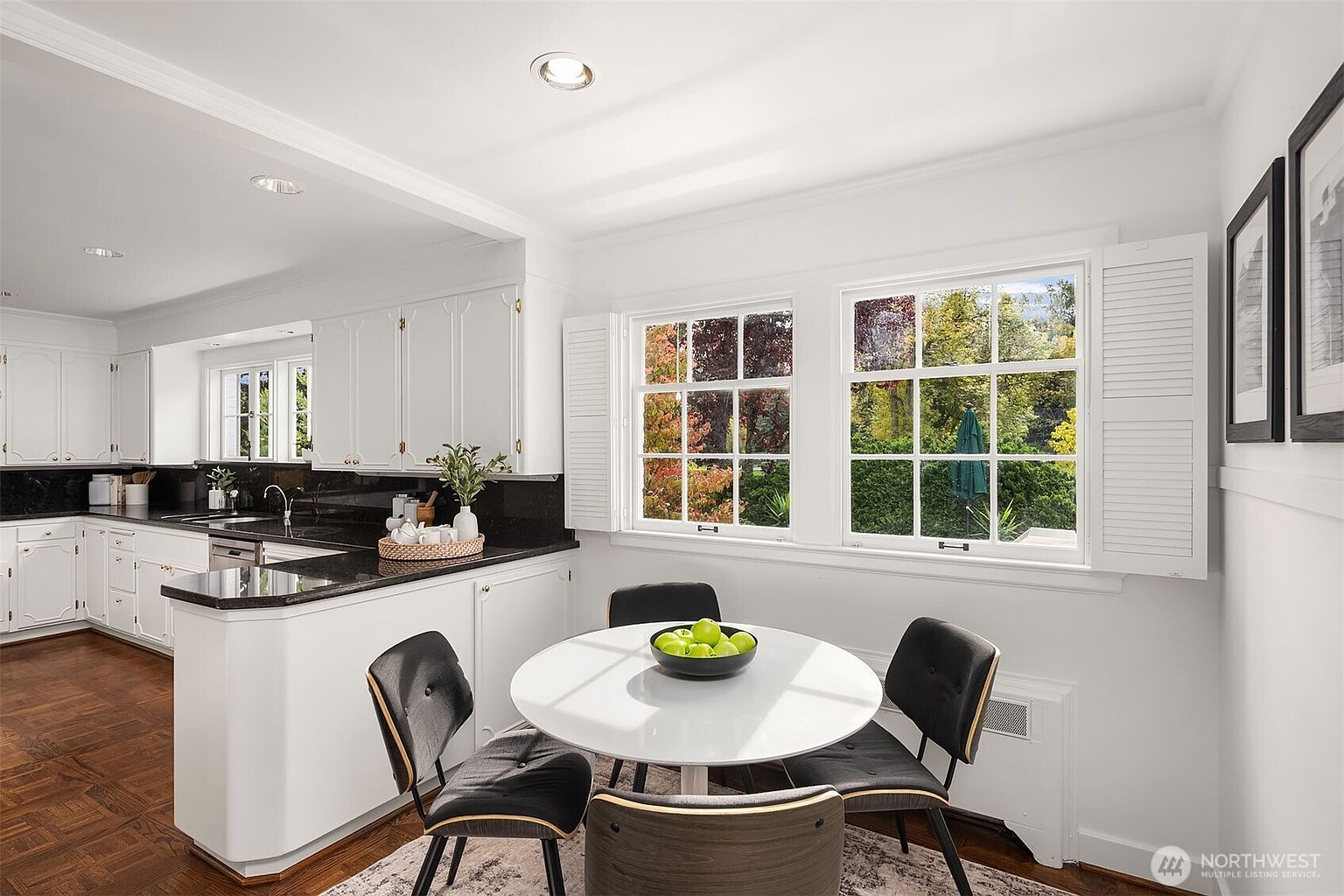 This is a bright and airy kitchen and dining area featuring white cabinetry, black countertops, and hardwood floors. A round white table with four chairs sits near a window offering a view of the outdoors. The space is well-lit and appears clean and modern, perfect for a cozy breakfast nook.