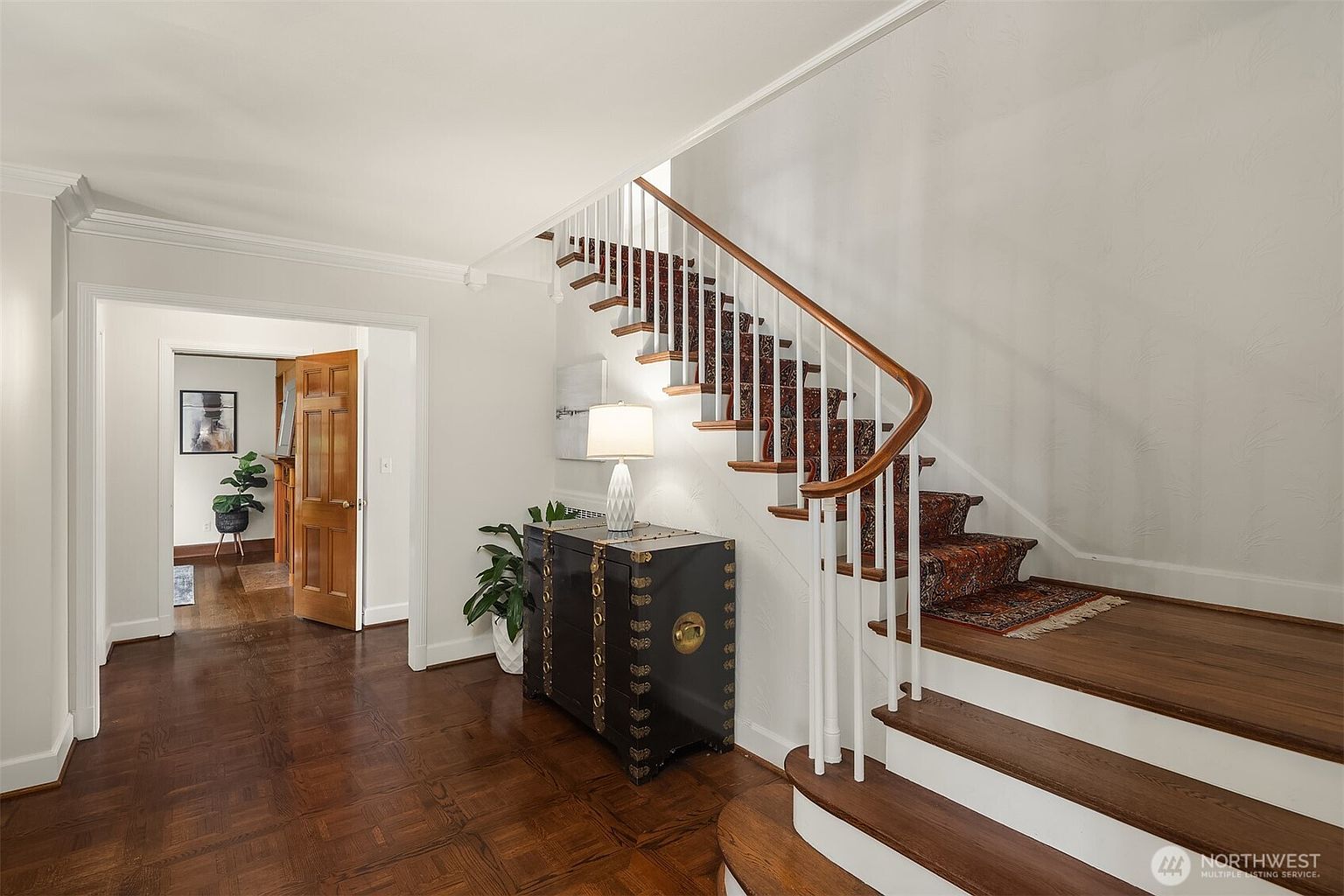 This interior shot showcases a well-lit hallway with hardwood floors and a staircase. The staircase features a patterned runner and white railings, adding a touch of elegance. A dark chest with brass accents sits against the wall, complemented by a table lamp and a potted plant, creating a welcoming and sophisticated entryway.