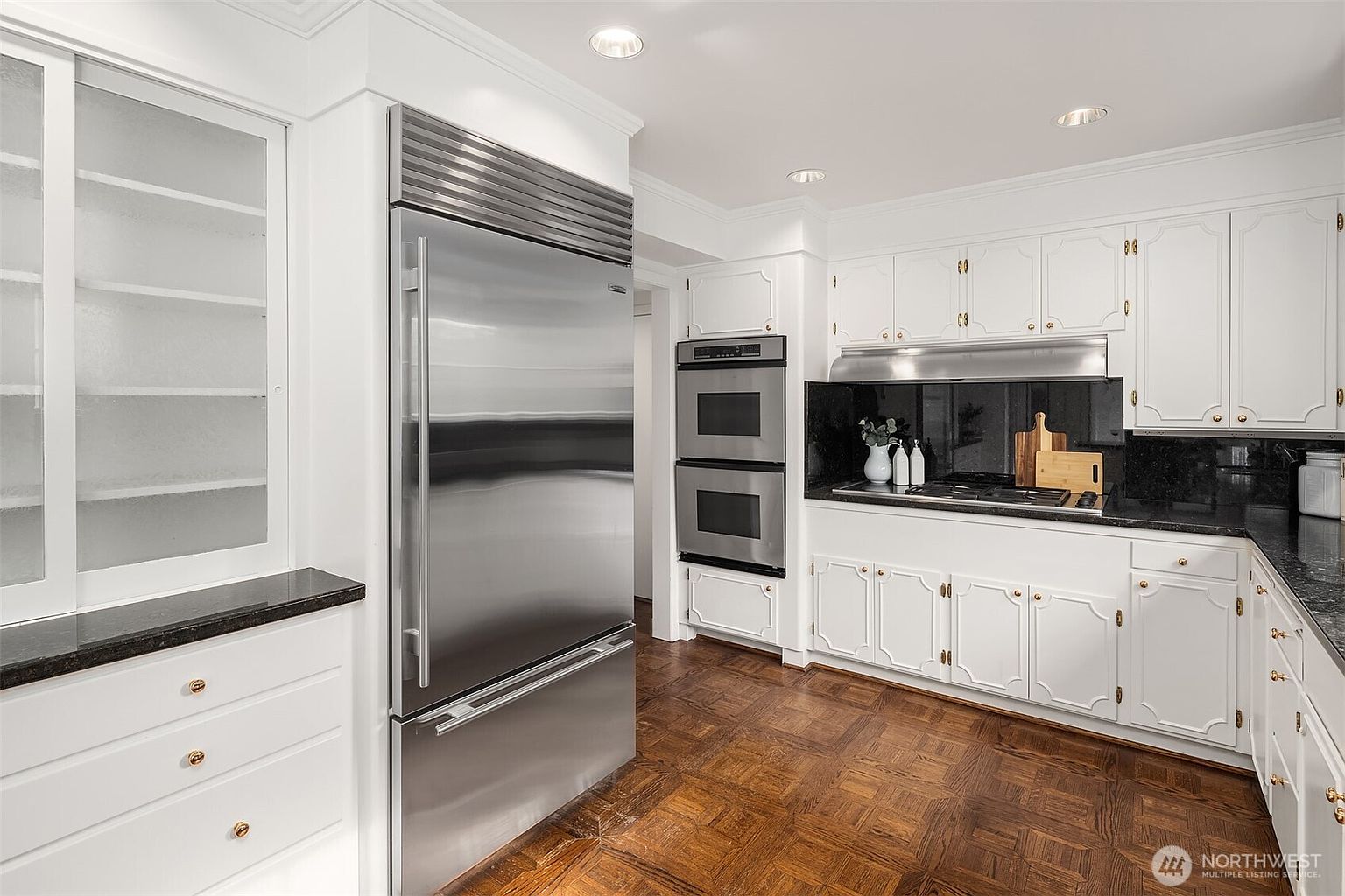This is a well-lit kitchen featuring white cabinetry with gold hardware, stainless steel appliances, and dark countertops. The floor is a classic parquet pattern, adding warmth to the space. The kitchen appears clean and functional, with a traditional yet timeless design.