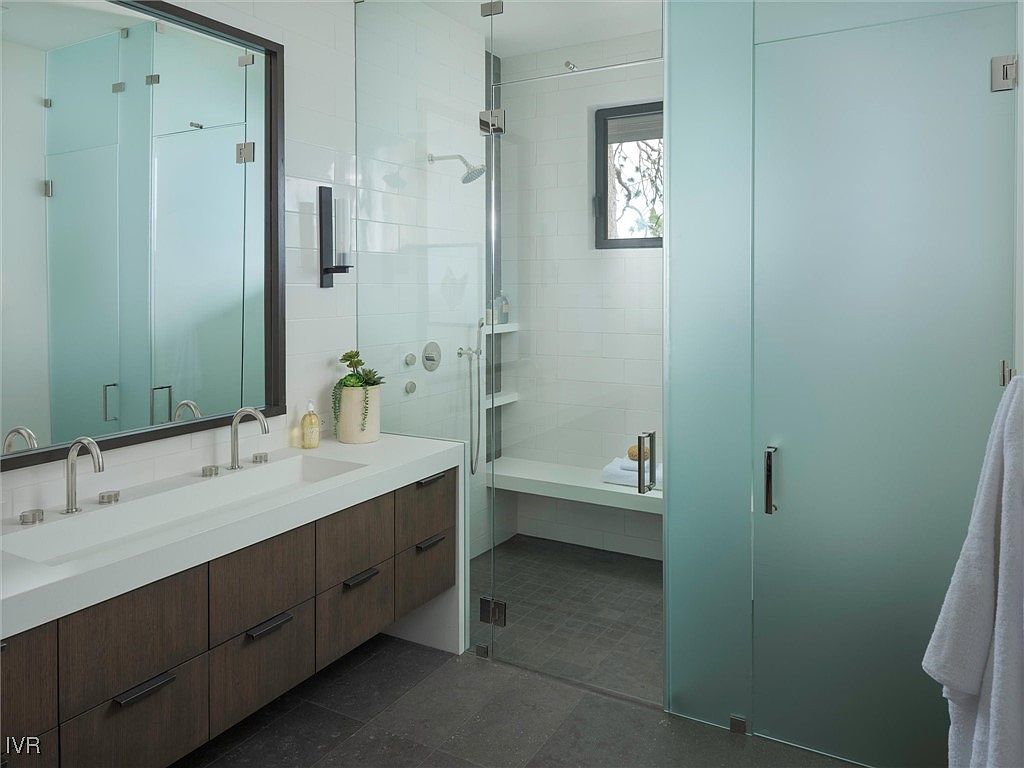 This is a modern primary bathroom featuring a double vanity with dark wood cabinets and a white countertop. A large mirror hangs above the vanity, and a glass-enclosed shower with a built-in bench is visible. The bathroom has a clean and minimalist design with white subway tile and dark gray flooring.