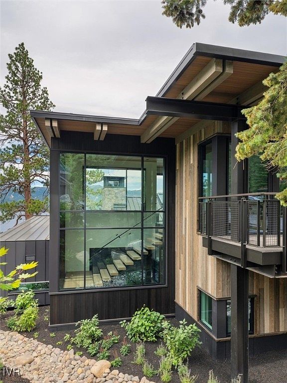 This is a side view of a modern house with a unique architectural design. The exterior features a combination of wood siding, dark metal accents, and large windows that offer a glimpse of the interior staircase. A small balcony extends from the side of the house, and the landscaping includes rocks and various plants.