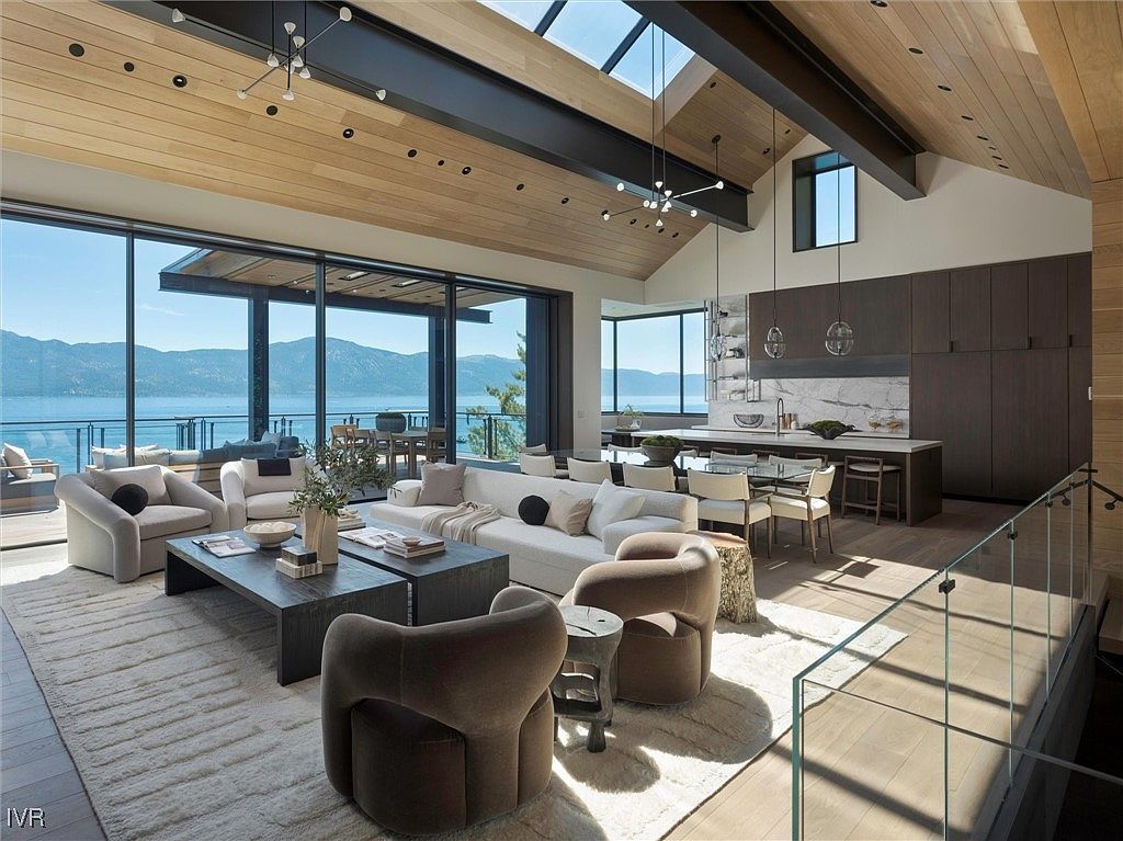 This is a spacious and luxurious living room with floor-to-ceiling windows offering stunning views of a lake and mountains. The room features a modern design with a high, wood-paneled ceiling, contemporary lighting fixtures, and comfortable seating arrangements. The open-concept layout seamlessly connects the living area to the kitchen and dining space, creating an inviting and sophisticated atmosphere.