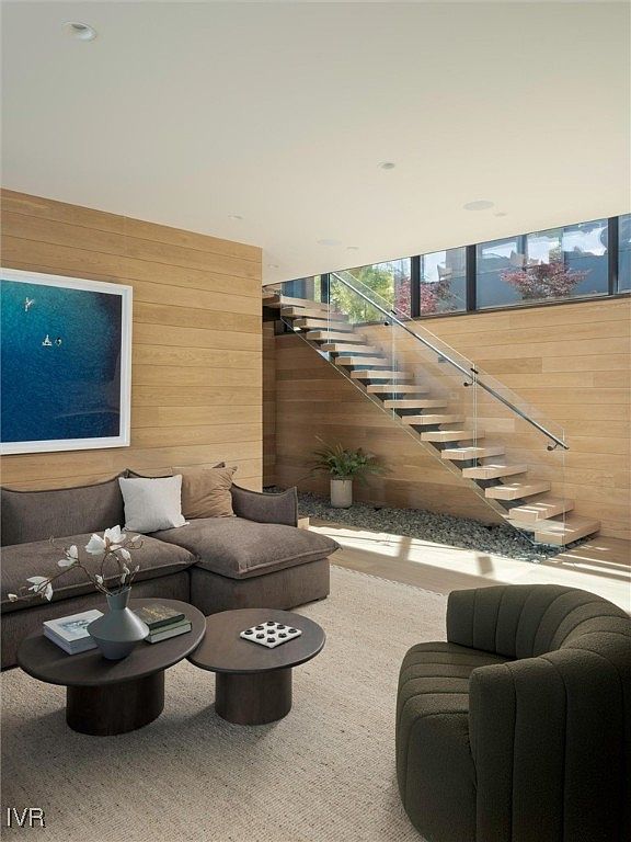 This is a stylish living room featuring a comfortable brown sectional sofa with decorative pillows, complemented by two round coffee tables. A modern armchair adds to the seating arrangement. The room is characterized by its clean lines, natural wood paneling, and a floating staircase with glass railings, creating a bright and airy atmosphere. A large framed artwork adds a touch of color to the space.