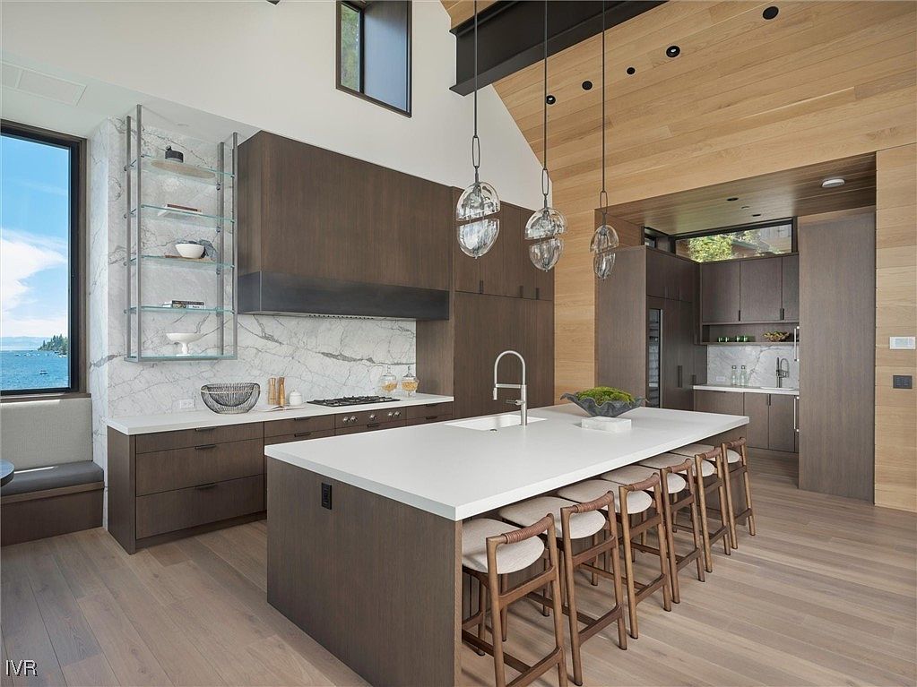 This is a modern kitchen with dark wood cabinetry, a white countertop island with seating, and stainless steel appliances. The kitchen features a marble backsplash and open shelving, with pendant lighting hanging above the island. The overall impression is one of luxury and sophistication.