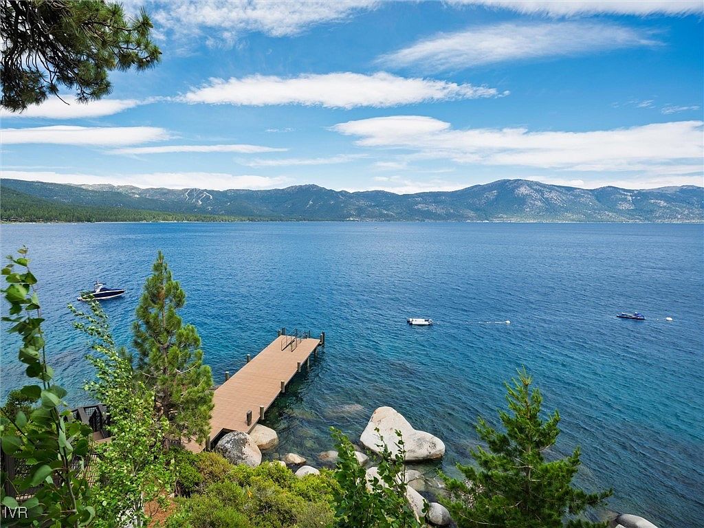This image showcases a stunning waterfront property featuring a private dock extending into a serene blue lake. The scene is framed by lush greenery and mature trees, with distant mountains providing a picturesque backdrop. Several boats are visible on the water, enhancing the sense of tranquility and recreational opportunities.