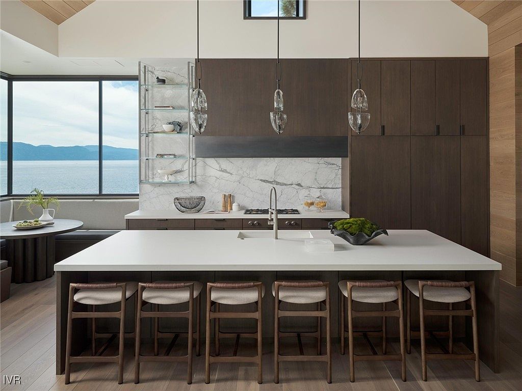 This is a modern kitchen with dark wood cabinetry and a large white countertop island with seating for six. The kitchen features pendant lighting, stainless steel appliances, and a large window with a view of a lake and mountains. The overall style is sleek and contemporary with a focus on natural light and views.
