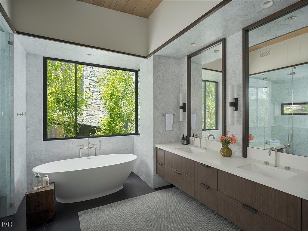 This is a luxurious primary bathroom featuring a freestanding soaking tub positioned beneath a large window offering a view of lush greenery. The bathroom boasts a double vanity with modern fixtures, complemented by large mirrors and elegant lighting. The space is finished with marble-like tiling and a neutral color palette, creating a serene and spa-like atmosphere.