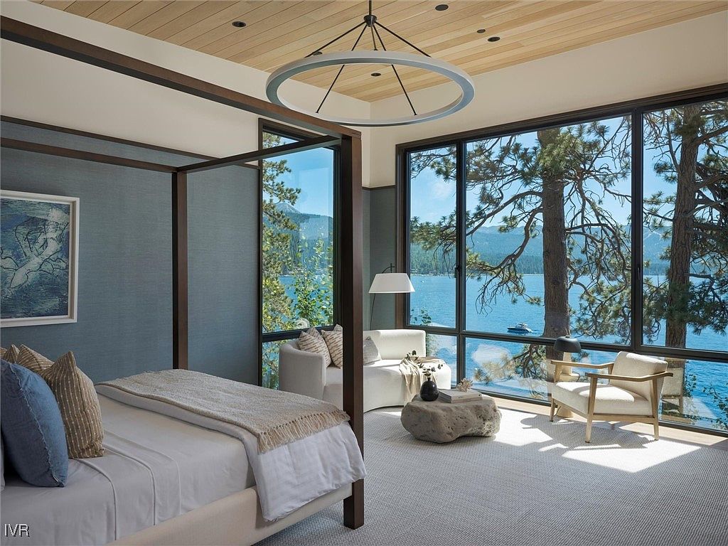 This is a luxurious primary bedroom featuring a four-poster bed with neutral linens and a textured blue headboard wall. Large windows offer stunning views of a lake and surrounding trees, creating a serene and inviting atmosphere. The room is furnished with modern seating and a unique stone coffee table, complemented by a contemporary circular chandelier.