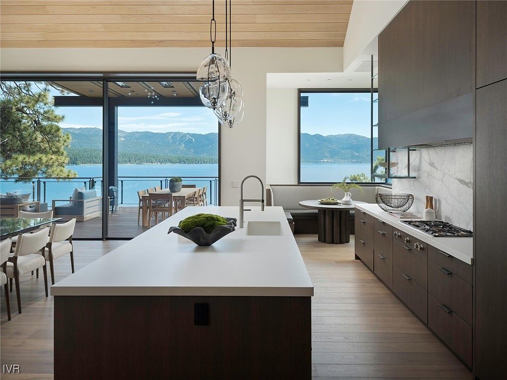 This is a modern kitchen with dark wood cabinetry and a large white countertop island. The kitchen features a view of a lake and mountains through large windows and sliding glass doors that lead to a deck. The overall impression is luxurious and serene, highlighting the connection to the natural surroundings.