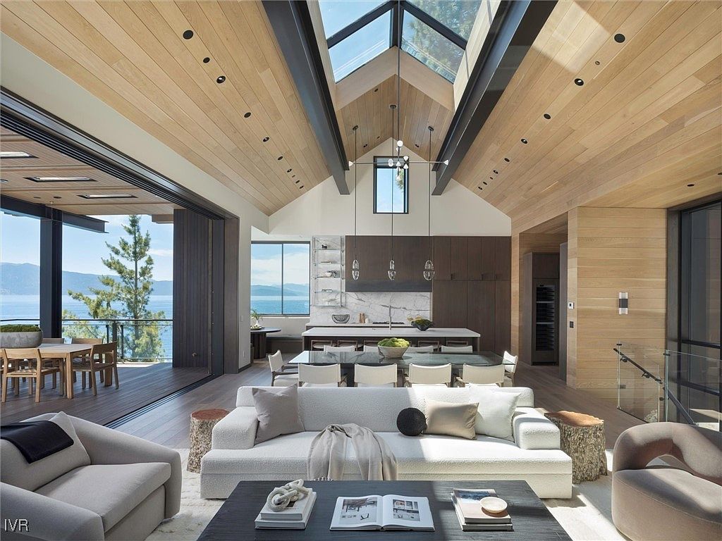 This is an interior shot of a luxurious living room that seamlessly blends with the kitchen and dining area. The open-concept space features a high, vaulted ceiling with wooden planks and skylights, allowing ample natural light. A large sliding glass door opens to a deck with stunning lake views, enhancing the indoor-outdoor living experience. The furniture is modern and comfortable, with a neutral color palette that complements the natural wood tones throughout the space.